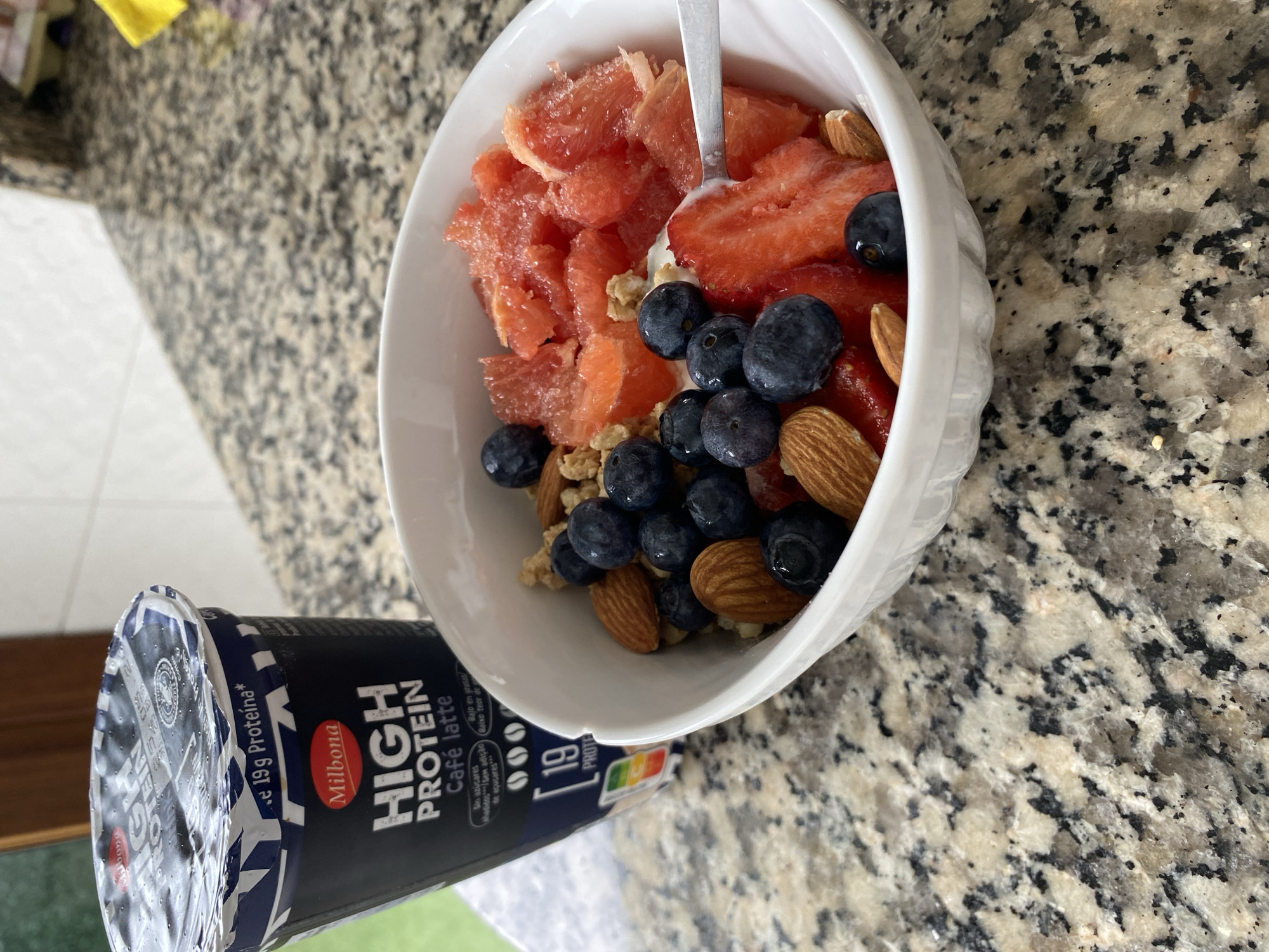 The image shows a bowl of fruit, nuts, and cereal on a granite countertop, next to a Milbona High Protein Café latte yogurt container. The bowl contains sliced strawberries, blueberries, grapefruit chunks, and almonds. A spoon rests in the bowl. The location is likely a home kitchen, but it is not possible to confirm that it is located in Gijón/Xixón, Spain.
