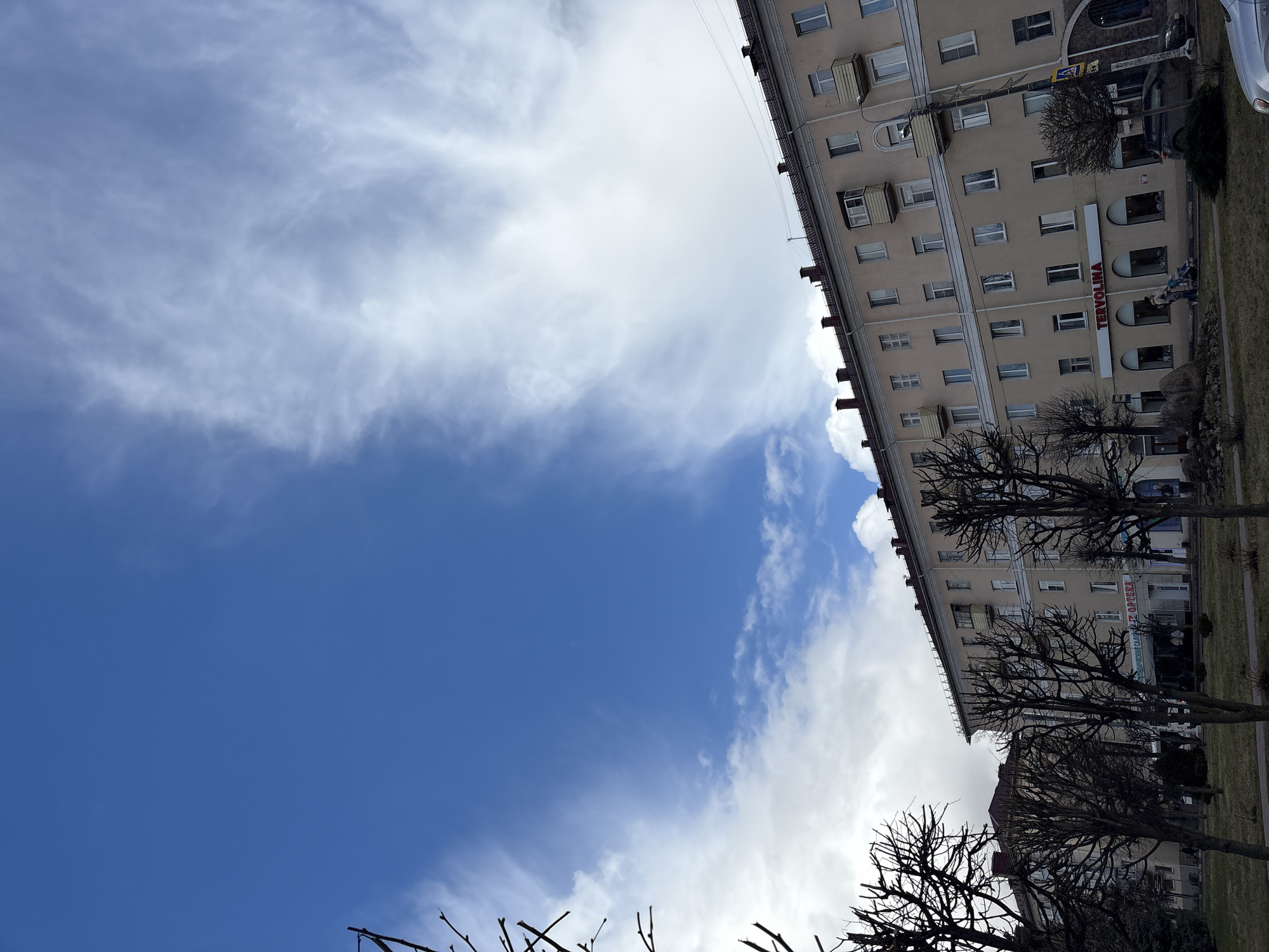 The image shows a view of a building in Klin, Russia. The building has a beige facade with multiple windows and balconies. A sign with the word "TERVOLINA" is visible on the building. Several bare trees are in front of the building. The sky is blue with white clouds.