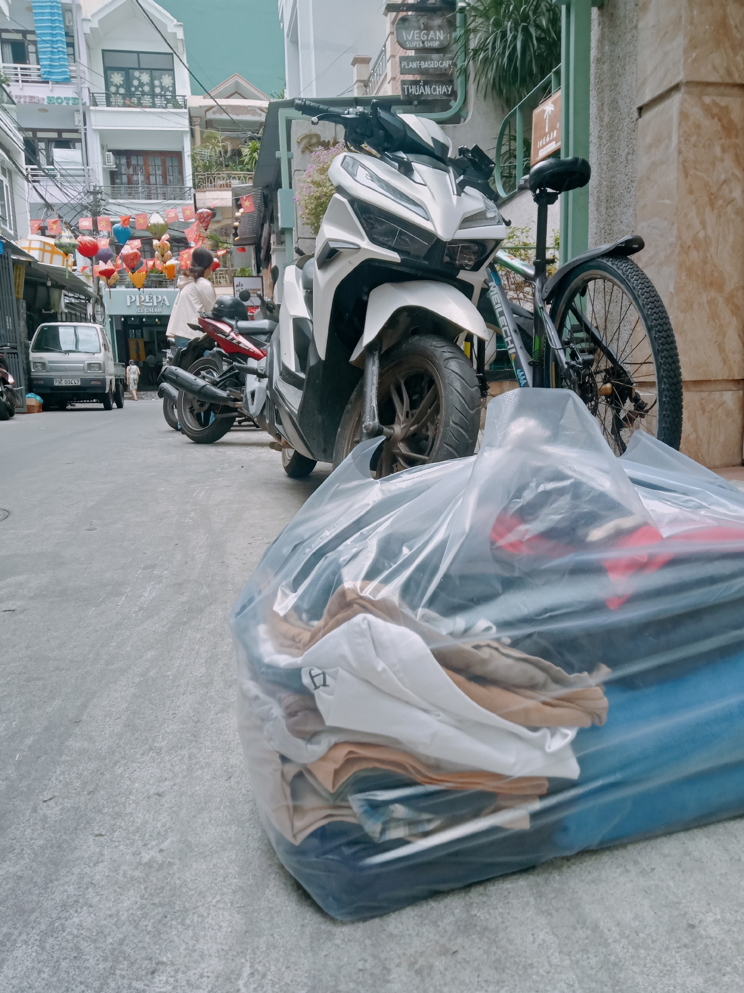 The scene shows a street view in Nha Trang Ward, Vietnam, cluttered with vehicles and objects. In the foreground, a large, clear plastic bag filled with clothing in various colors, including white, tan, and blue, dominates the lower portion of the frame. Behind the bag, a white scooter is parked, partially obscuring a bicycle with the brand name "MEILECHI" visible on its frame. Further down the street, there's a red motorcycle parked behind the scooter.
In the background, a white van with the license plate "79C 00447" is visible, parked in front of a building labeled "PRÉPA ICE CREAM". A woman wearing a white top stands near the ice cream shop.
The street is lined with buildings, one of which has a sign that reads "TIELY HOTEL". A vertical sign advertises a "VEGAN SUPER SHOP" and a "PLANT-BASED CAFE THUẨN CHAY". A string of colorful lanterns is strung across the street. The environment appears urban and crowded.