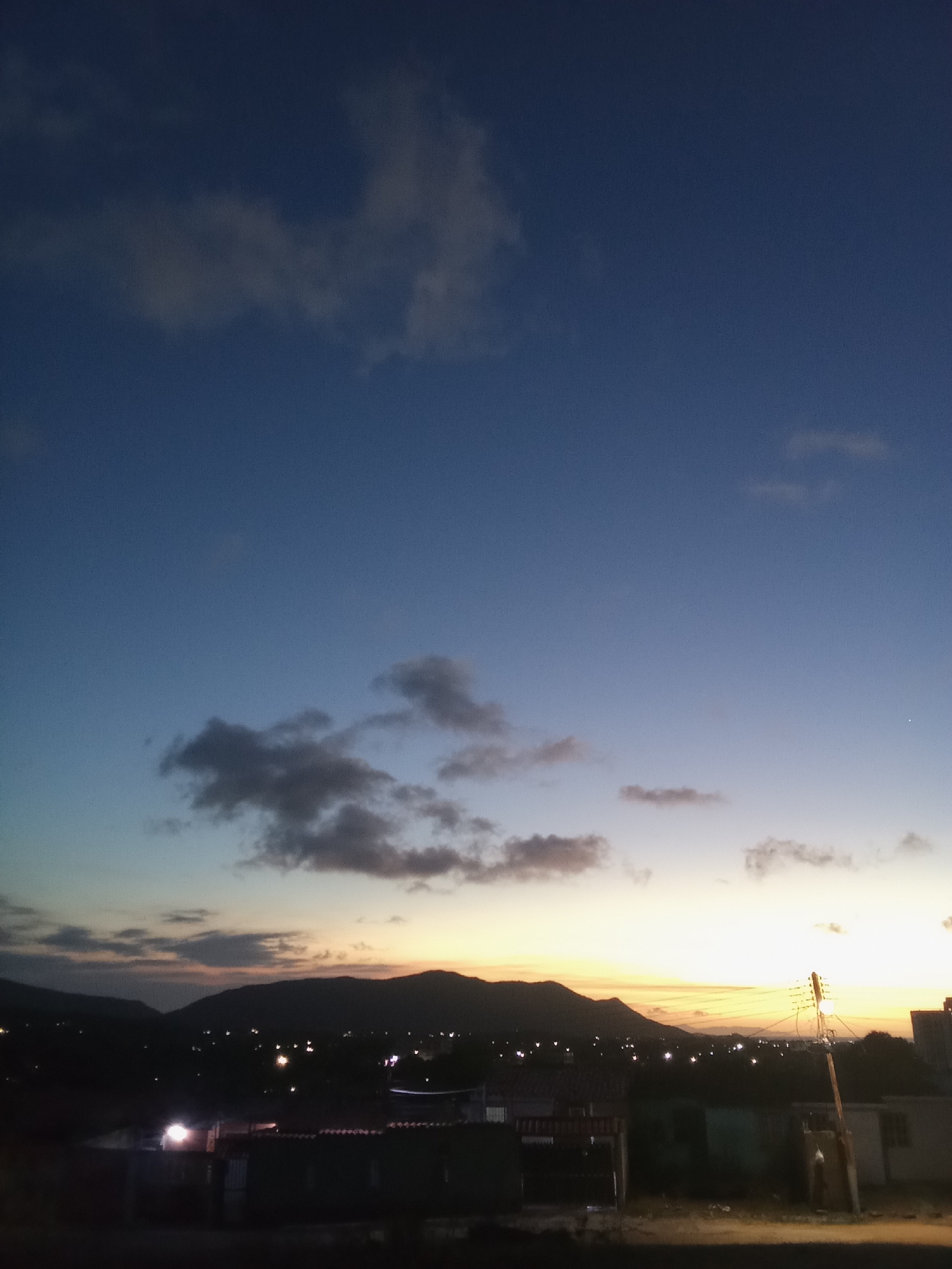 An image depicting the horizon, taken at twilight in or near Juan Griego, Venezuela. In the middle ground, there is a chain of mountains. In the foreground is residential housing. The sky transitions from a deep blue color above to a brighter yellow and orange hue near the horizon. There are scattered clouds visible in the sky. Streetlights and lights from homes dot the landscape. A utility pole with electrical wires stands to the right.