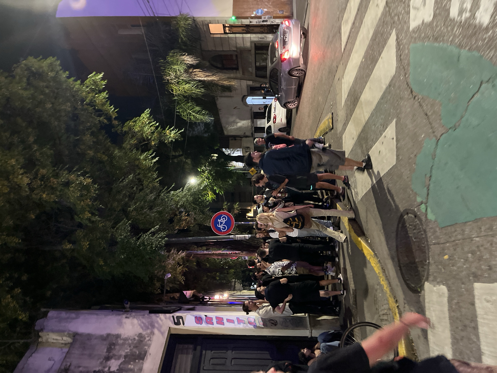 The image shows a nighttime scene on a street in Buenos Aires, Argentina. A group of people are standing on a crosswalk. Several parked cars are visible. There's a bicycle lane painted green on the street. A building with a sign above the door is on the left side of the street. A bicycle lane sign is visible on a pole.