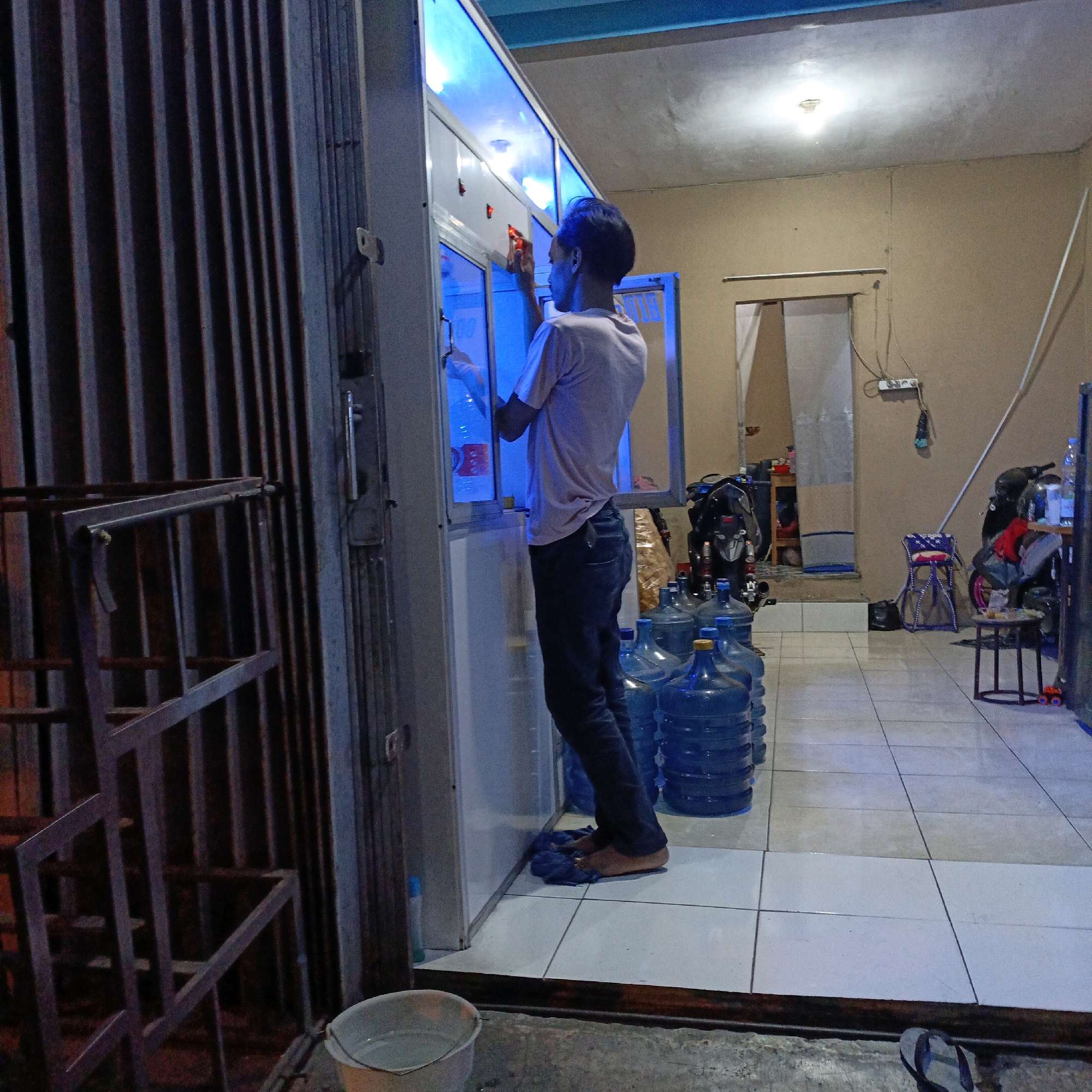 The image depicts the interior of a small shop in Salatiga, Indonesia, likely a water refill station or a convenience store. In the foreground, a person is standing, facing a glass-fronted cabinet illuminated by bright blue LED lights. They are wearing a light-colored t-shirt and dark trousers, with dark flip-flops on their feet. The person appears to be reaching into the cabinet, possibly attending to goods or services offered within.
Several large, blue plastic water jugs are stacked in front of the cabinet, indicating the shop's primary business. The floor is tiled in a white and light gray pattern. To the left, a dark metal grate structure is visible, possibly part of the shop's entrance or display. In the background, a motorcycle is parked, and other shelves or cabinets are present, suggesting a cluttered storage or retail space. The walls are a neutral, sandy color. The overall lighting is a mix of the bright blue from the cabinet and the warmer glow of an overhead fluorescent light. The scene suggests a late evening or nighttime operation.
