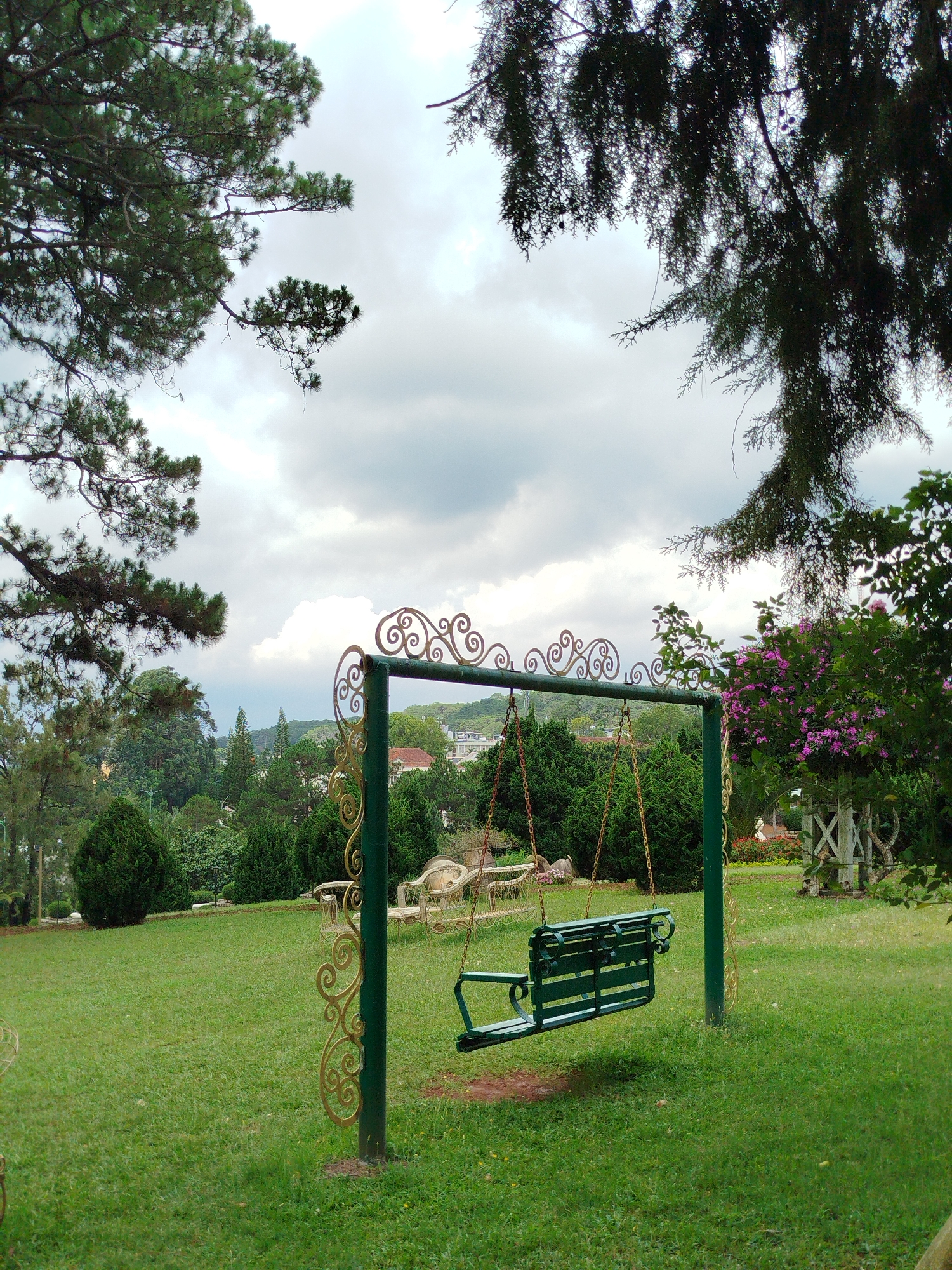 The image depicts a green metal swing set with ornate gold-colored scrollwork in a grassy park or garden setting. The swing is suspended by chains. The grass is green and appears well-maintained. In the background, there is a variety of shrubs and trees, with glimpses of buildings indicating an urban or suburban area. The sky is overcast with grey clouds. The location is in Da Lat, Vietnam. The swing set is positioned within a landscaped area, possibly a public park or a garden associated with a hotel or resort. There are benches and decorative garden structures visible in the background. The scene is tranquil and suggestive of a recreational space.