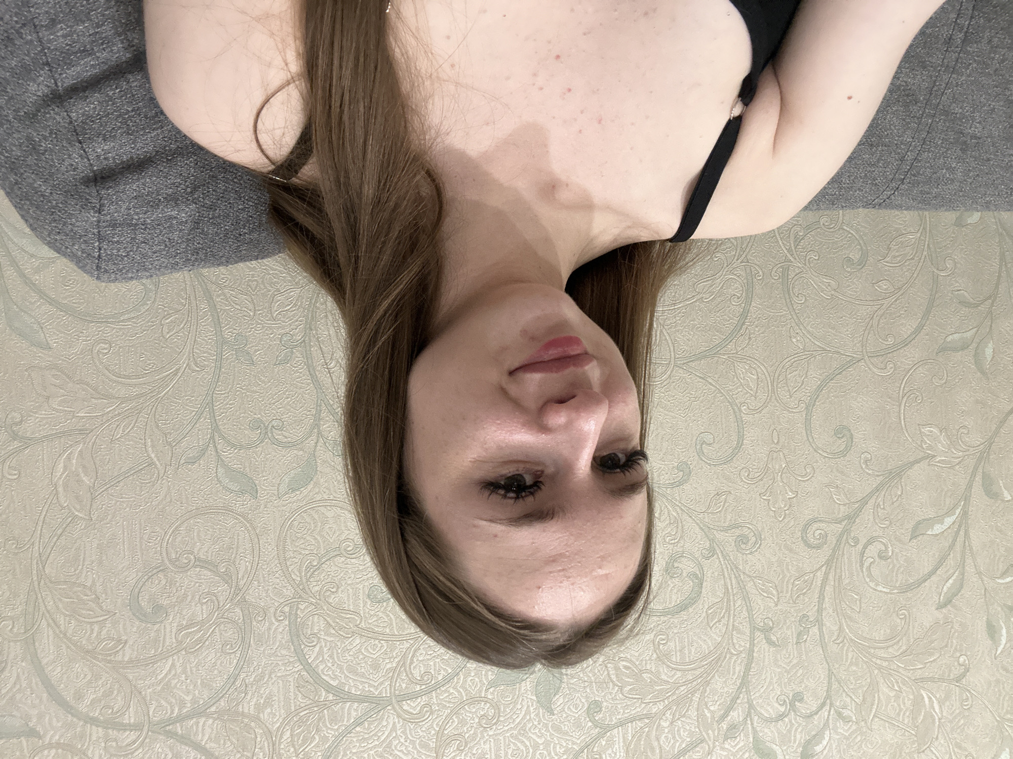 A young woman with long, light brown hair is shown from the chest up, looking slightly to her left. She is wearing a black top. Her skin has some blemishes. She is sitting on a gray couch in front of a wall with a floral wallpaper. The scene is indoors, likely a room in a residential setting.