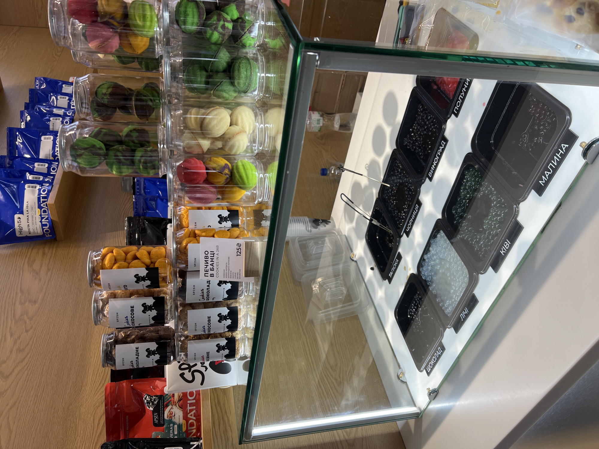 The image displays a retail counter in a shop or cafe, likely located in Odesa, Ukraine. The scene features two main sections: a vertical display of packaged goods and a glass-enclosed counter showcasing various toppings.
On the left, several clear plastic jars are stacked, filled with colorful, shell-shaped cookies or pastries, including green striped, cream, and multi-colored varieties. Below these, more jars contain "SPESH" brand cookies, labeled as "шоколадне" (chocolate), "кокосове" (coconut), and "всяне" (oat). One jar prominently features "ПЕЧИВО В БАНЦІ COOKIES IN A JAR" with a price of "125₴". Bags of coffee or similar products are also visible, some labeled "FOUNDATION," "KOCTA PIKA LA ISLA," and "APSEN COFFEE ROASTERS."
The right side of the image reveals a glass display case with black square containers filled with small, spherical toppings. Each container is labeled in Ukrainian: "ПОЛУНИ" (Strawberries), "МАЛИНА" (Raspberry), "ВИНОГРАД" (Grapes), "ЧОРНИЦЯ" (Blueberry), "ТАПІОКА" (Tapioca), "КІВІ" (Kiwi), "ШОКОЛАД" (Chocolate), and "ЛІЧІ" (Lychee). The toppings vary in color and texture, from red and dark to clear and green. Reflections in the glass show water bottles and other elements of the interior setting.
The overall impression is of a modern establishment offering a selection of pre-packaged baked goods and a range of customizable toppings, possibly for beverages like bubble tea or desserts. No people or specific time of day are discernible.