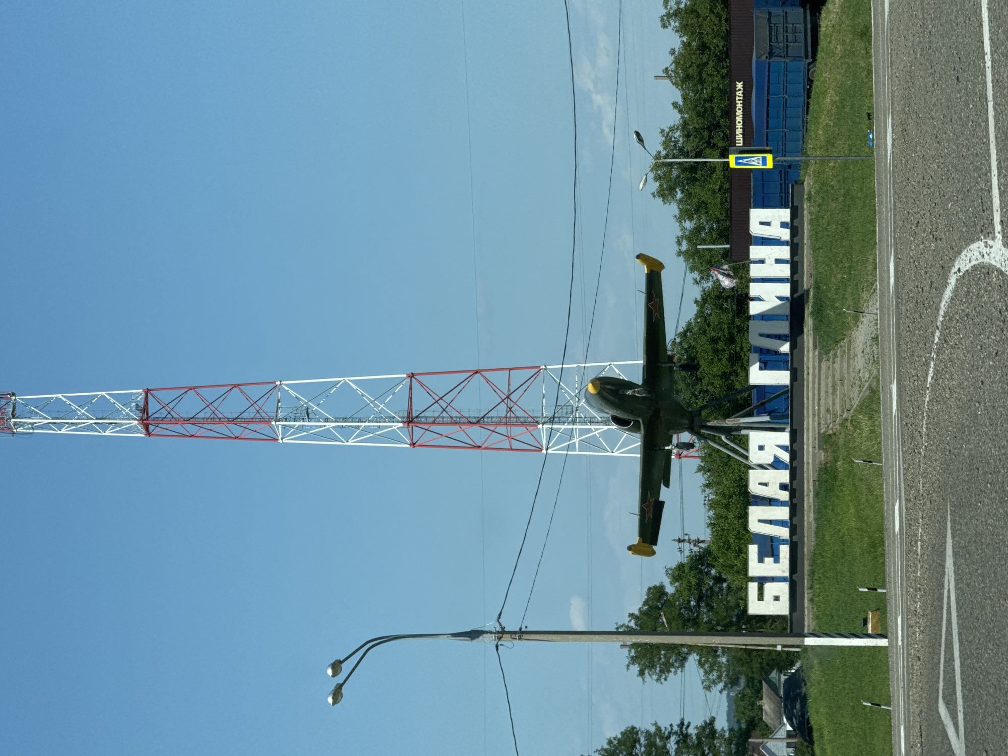 The image depicts an outdoor scene in the city of Belaya Glina, Russia, featuring a prominent telecommunications tower in the background and a monument in the foreground. The monument consists of a vintage military jet airplane mounted on a stand, with large white Cyrillic letters spelling "БЕЛАЯ ГЛИНА" (Belaya Glina) positioned below it. A lamppost with two lights stands to the left of the monument. In the far right background, a blue truck trailer is visible, along with a sign that appears to read "ШИННОМОНТАЖ" (Tire fitting) and a yellow pedestrian crossing sign. The environment is characterized by a clear blue sky, green grass, and trees. The image is taken from a low angle, looking upwards towards the tower and monument.