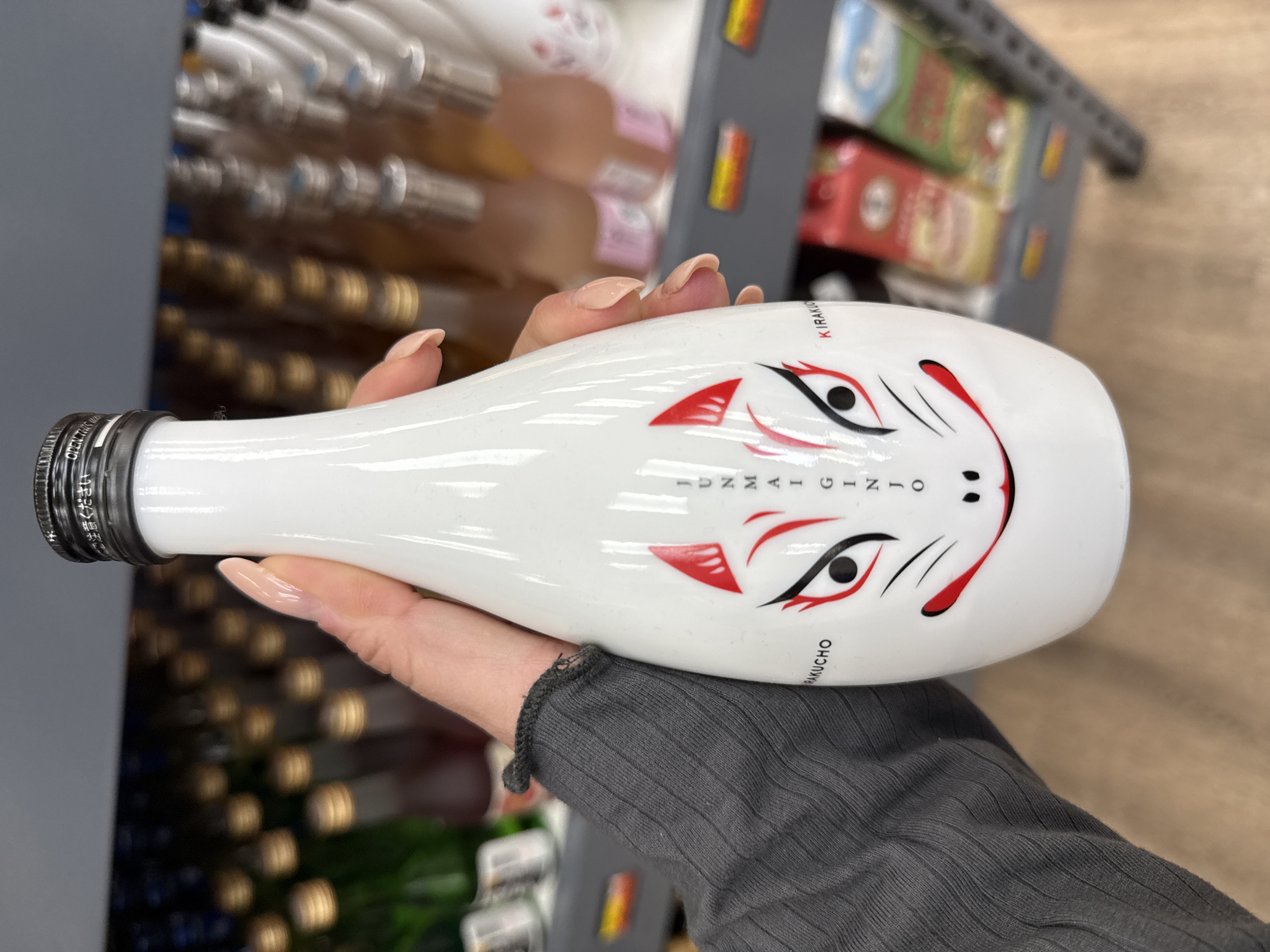A person is holding a white bottle with a black and red fox-like design in what appears to be a retail store in Davie, United States of America. The bottle has a metallic cap. The person is wearing a grey, long-sleeved shirt. Other bottles and items are visible on shelves in the background, slightly out of focus.