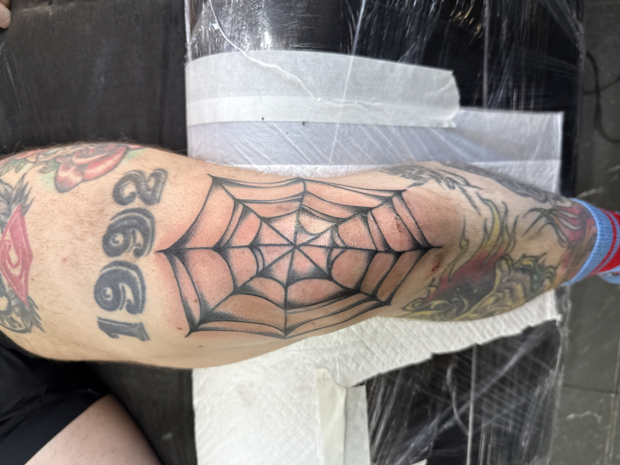 An adult's arm, heavily tattooed, rests on a surface partially covered with white paper towels and clear plastic wrap. The arm is tan with dark body hair. The tattoos include a large black spiderweb centered around the elbow, the number "1992," and other various designs with red, yellow, and black ink. The arm is positioned horizontally across the frame. The location in Moscow, Russia, cannot be determined from the image.
