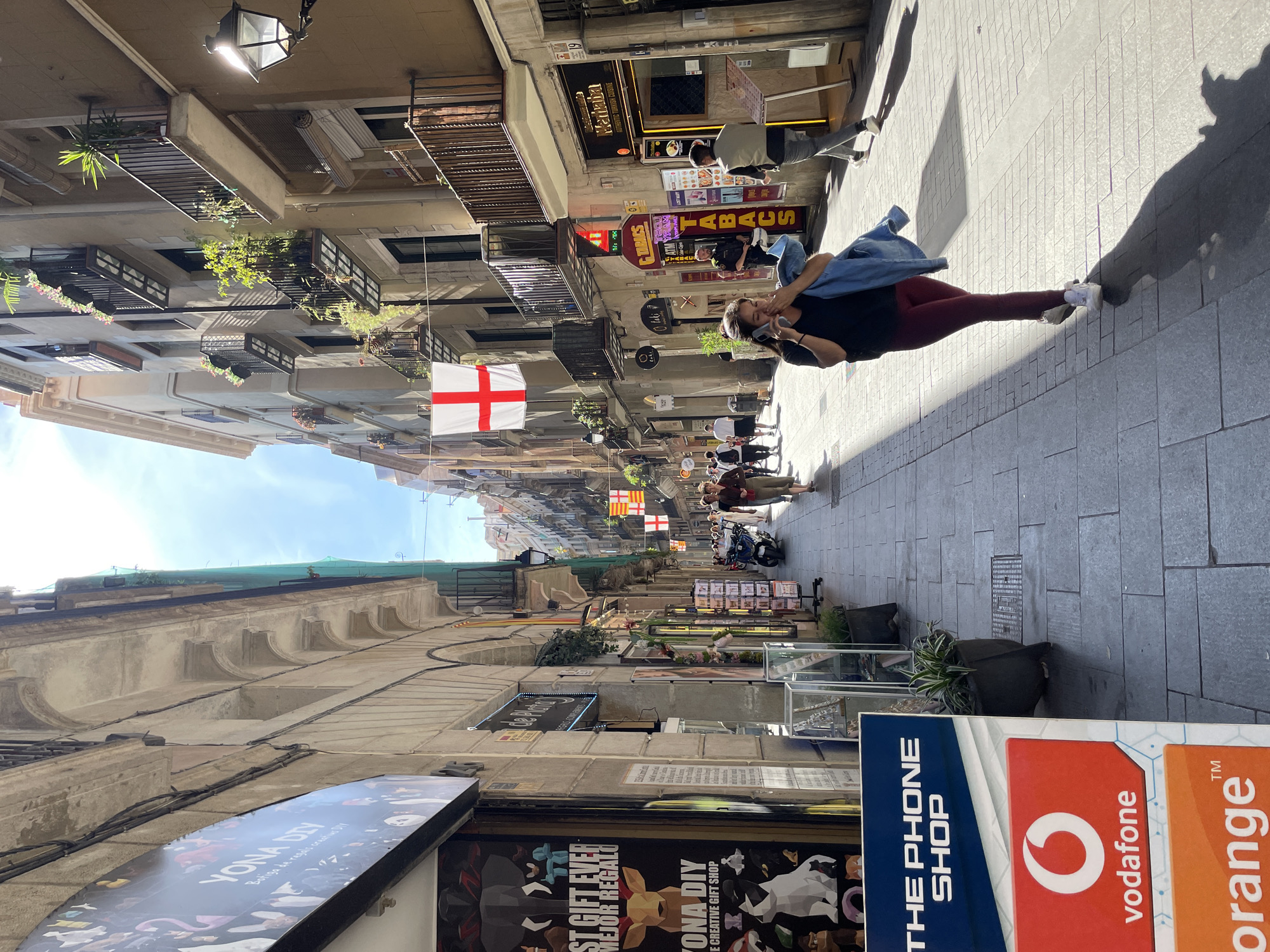 A street view in Barcelona, Spain. A woman wearing dark leggings, a dark shirt, and white shoes is walking down a street. She is carrying a denim jacket draped over her shoulder. There are several flags with red crosses on a white background, as well as flags with yellow and red stripes, hanging above the street. There are shops on both sides of the street with signs in both English and Spanish, including "THE PHONE SHOP," "vodafone," "orange," "YONA DIY," "Merhaba," and "TABACS." There are also several people walking down the street. Buildings on either side of the street feature balconies with greenery.