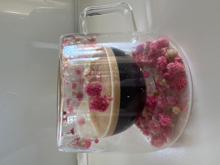 A clear double-walled glass mug, containing a dark liquid topped with a light brown crema, identifiable as an espresso, rests on a light-colored surface. The inner chamber of the mug holds the coffee. The void between the mug's inner and outer glass walls, including the base of the outer chamber, is filled with numerous small, dried flowers; these are predominantly vibrant pink, interspersed with some lighter pink or off-white elements. The mug features an integrated clear glass handle on its right side. The background is a plain, light-toned, indistinct surface. The scene is situated in Varna, Bulgaria.