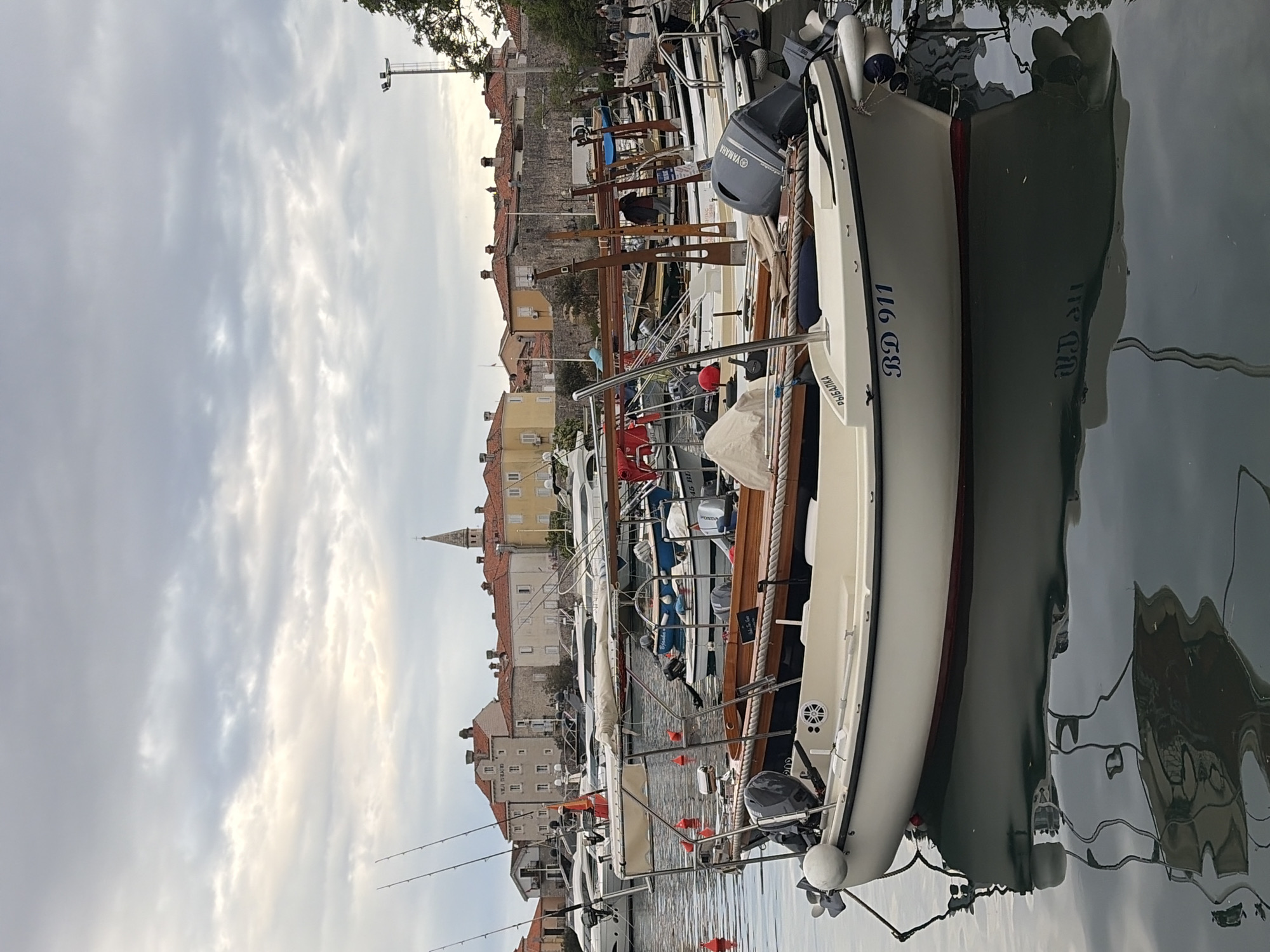 The image depicts a harbor scene in Budva, Montenegro, with a focus on several docked boats. In the foreground, a white boat with the identification number "BD 911" is prominently featured, partially obscuring the view of other boats behind it. These boats are moored along a wooden dock. Beyond the boats, the cityscape of Budva rises, showcasing buildings with orange tiled roofs, a church spire, and stone walls. The sky is overcast with light gray clouds. The water reflects the boats and the cityscape, creating a mirror image effect. There are also fishing rods visible on one of the boats.