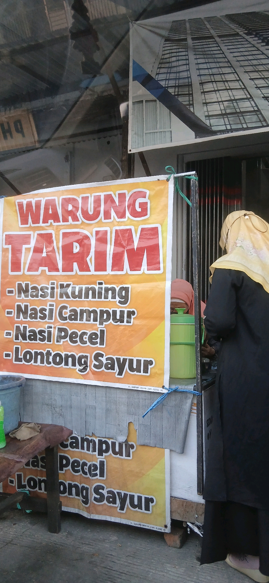 A banner for "WARUNG TARIM" is displayed in front of a building. The banner lists menu items: "- Nasi Kuning", "- Nasi Campur", "- Nasi Pecel", and "- Lontong Sayur". Two individuals are partially visible. One is wearing a light yellow hijab and a black abaya, standing near a green plastic container. Another person, wearing a light brown hijab, is partially visible behind the green container. The background shows a metal grate door and a partial view of a building facade with a large graphic of a skyscraper. The environment appears to be an outdoor food stall or small restaurant in Samarinda, Indonesia, judging by the provided context. The banner is affixed to wooden poles and secured with blue and green strings. A trash bin is visible on the left side of the image.