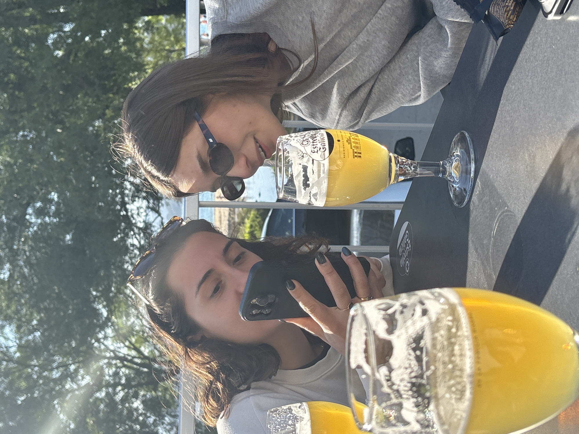 Two women sit at a table outdoors, likely on a patio, in O Porto do Son, Spain. The woman in the foreground, wearing a white shirt and sunglasses on her head, uses a dark-colored mobile phone, her hand adorned with a ring and fingernails painted a dark color. A beer glass filled with golden liquid is prominently positioned in front of her. The second woman, wearing a gray hooded sweatshirt and sunglasses, is next to a similar beer glass. The table is dark, and there's sunlight creating shadows. A building is behind them. The background features green foliage, indicating trees and vegetation. The glasses contain "Estrella Galicia" beer.
