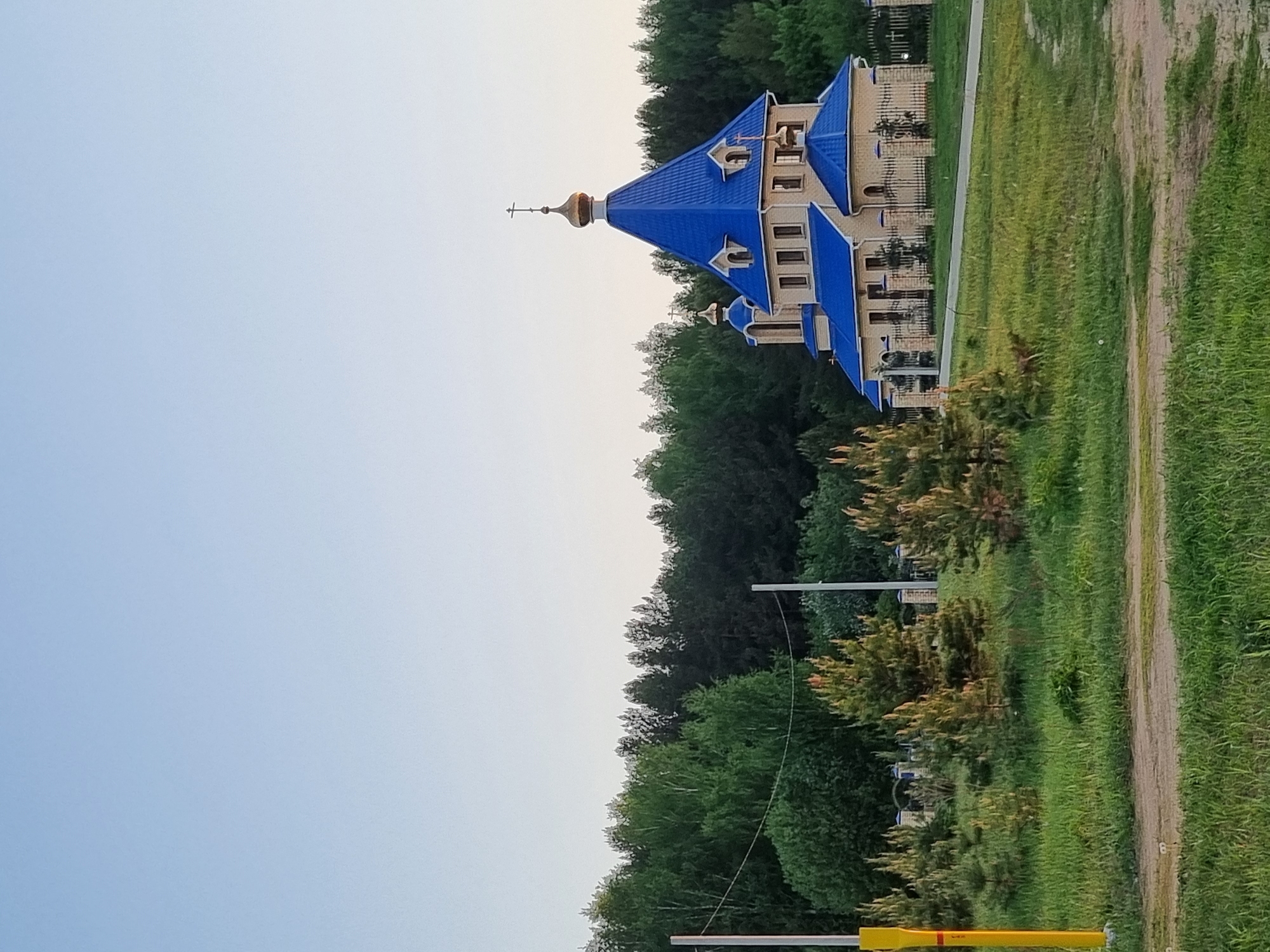 The image depicts a church with a distinctive blue roof located in Chaikovsky, Russia. The church stands amidst green vegetation, with a dirt path running alongside it. The sky is light blue, suggesting either early morning or late evening. The architecture of the church is traditional with a golden dome topped with a cross. There are trees and bushes surrounding the structure, indicating a natural environment.