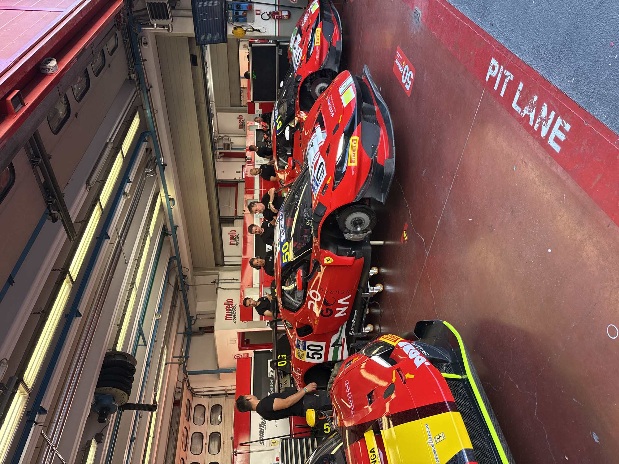 The image depicts a pit lane scene at the Mugello Circuit in Scarperia, Italy. Several high-performance racing cars, predominantly red with accents of yellow and black, are parked in a garage area. One car, a red Ferrari, is prominently positioned with the number "50" clearly visible on its side and front. Another red Ferrari race car with the number "51" is behind it. A partially visible red and yellow race car is in the foreground on the left.
Several individuals, presumably crew members, are present in the garage, some interacting with the cars and others observing. They are dressed in casual attire, with some wearing t-shirts.
The environment appears to be a professional racing facility. The garage is well-lit with fluorescent lighting fixtures overhead. Various pieces of equipment, including what appear to be tire stands and tool chests, are visible. Signs with "Mugello Circuit" branding are displayed on the walls. The floor of the pit lane is marked with red and white lines, including a "PIT LANE" designation. The overall impression is one of a bustling operational area within a racing circuit.