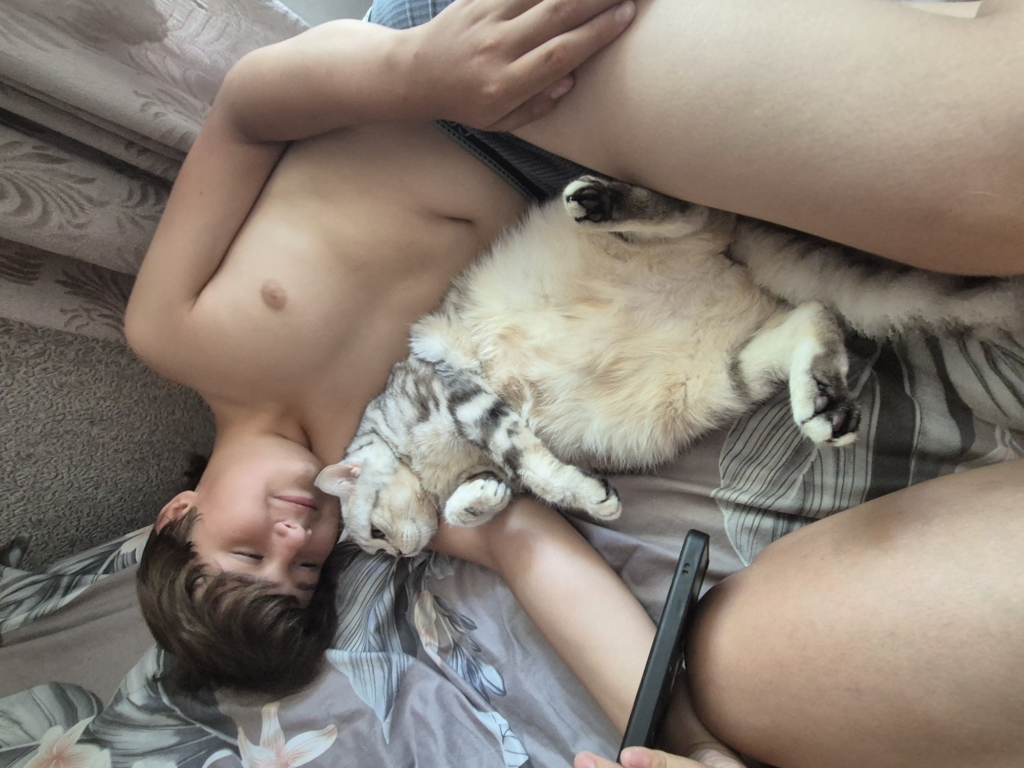 The image shows a close-up, overhead view of a young person lying on a bed with a cat. The person is lying on their back, with their head turned to the side, eyes closed, and a relaxed expression. They are shirtless. A light-colored cat with tabby markings is curled up on top of the person's chest and abdomen, also appearing to be sleeping or resting. One of the cat's paws is visible. A dark-colored smartphone is held by the person's hand, positioned near their chest.
The setting appears to be indoors, likely a bedroom, with bedding that has a floral pattern. The lighting suggests it might be daytime, possibly morning or afternoon, given the ambient light. The overall mood is one of comfort and relaxation. There is no visible text in the image. The location context is Sozonivka, Ukraine.