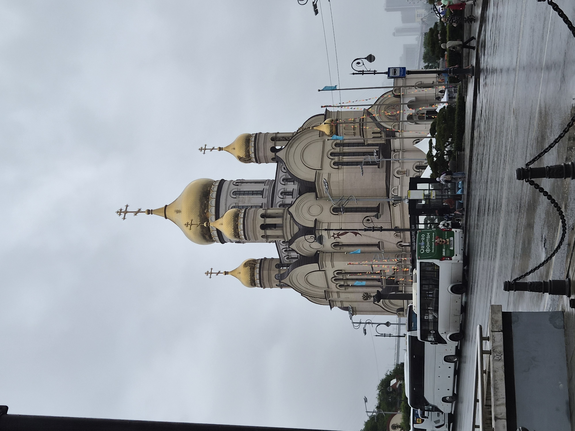 The image depicts a street scene in Vladivostok, Russia, featuring a large, ornate Orthodox church with multiple golden domes. The church is the central and dominant subject, with intricate architectural details. In the foreground, a wet street reflects the overcast sky, indicating recent rain. Several white tour buses are parked along the side of the road, suggesting a tourist area. Pedestrians are visible on the sidewalk, some walking and others standing near the buses or the church entrance. The overall atmosphere is subdued, likely due to the cloudy weather. A flagpole with a blue flag and decorative bunting are visible near the church. The city skyline can be seen faintly in the background, suggesting an urban setting.