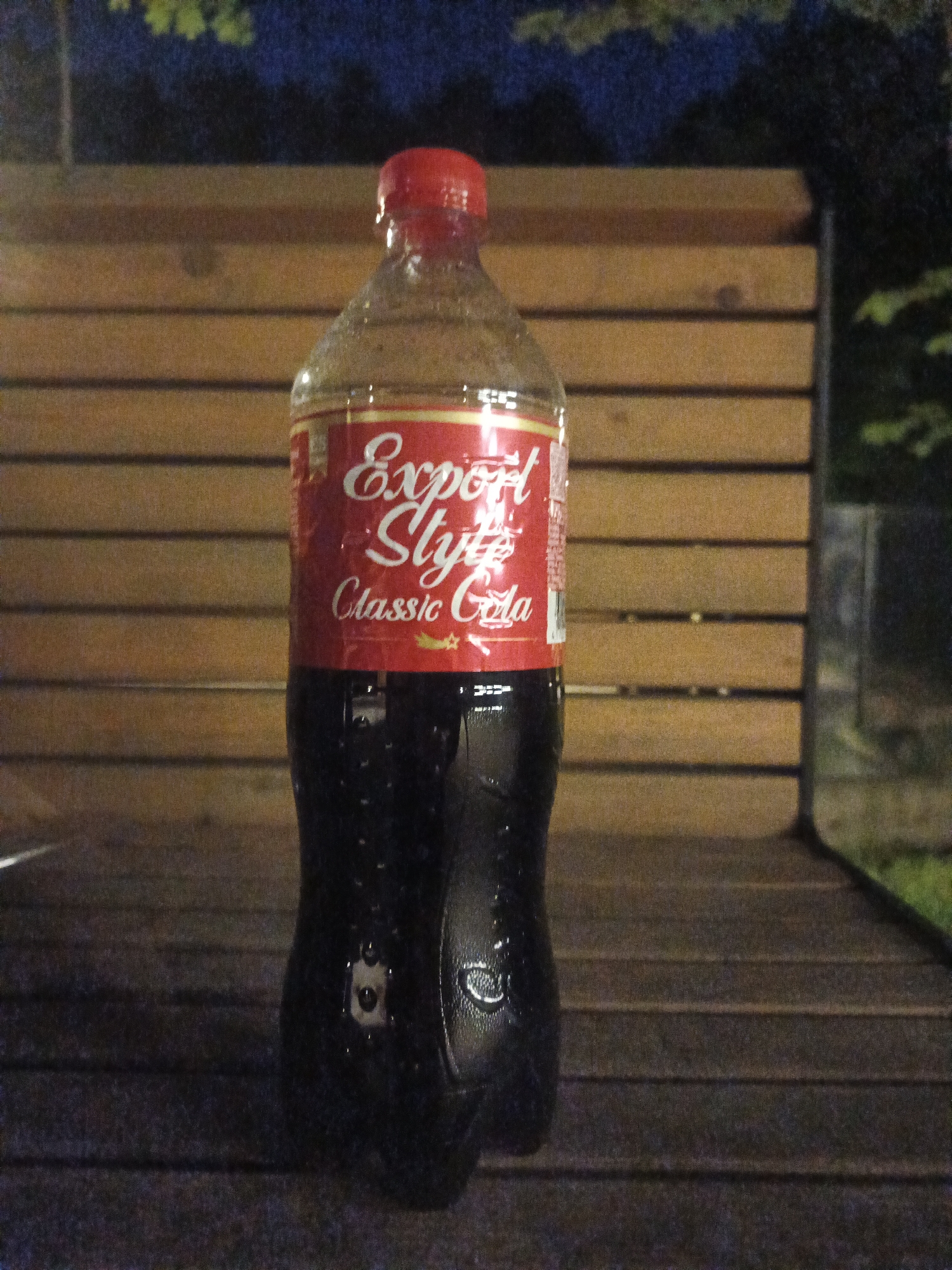 A medium shot captures a full, upright plastic bottle of "Export Style Classic Cola" against a backdrop of wooden planks. The bottle is mostly full, with the dark cola inside showing condensation droplets on the exterior, suggesting it's cold. The red cap is clearly visible at the top, and the label is predominantly red with white cursive text that reads "Export Style" and "Classic Cola." There's a small, stylized yellow star or emblem below the text. The bottle is positioned centrally on a wooden surface, possibly a bench or table, with the grain of the wood running horizontally. Behind the bottle, more wooden planks form a wall or barrier, also with horizontal grain. In the upper part of the image, blurred greenery and dark, indistinct shapes suggest foliage and trees, hinting at an outdoor setting. The lighting is dim, indicating it might be dusk or nighttime. The overall scene is still and quiet. No people are visible. The location context is Sof'ino, Russia.