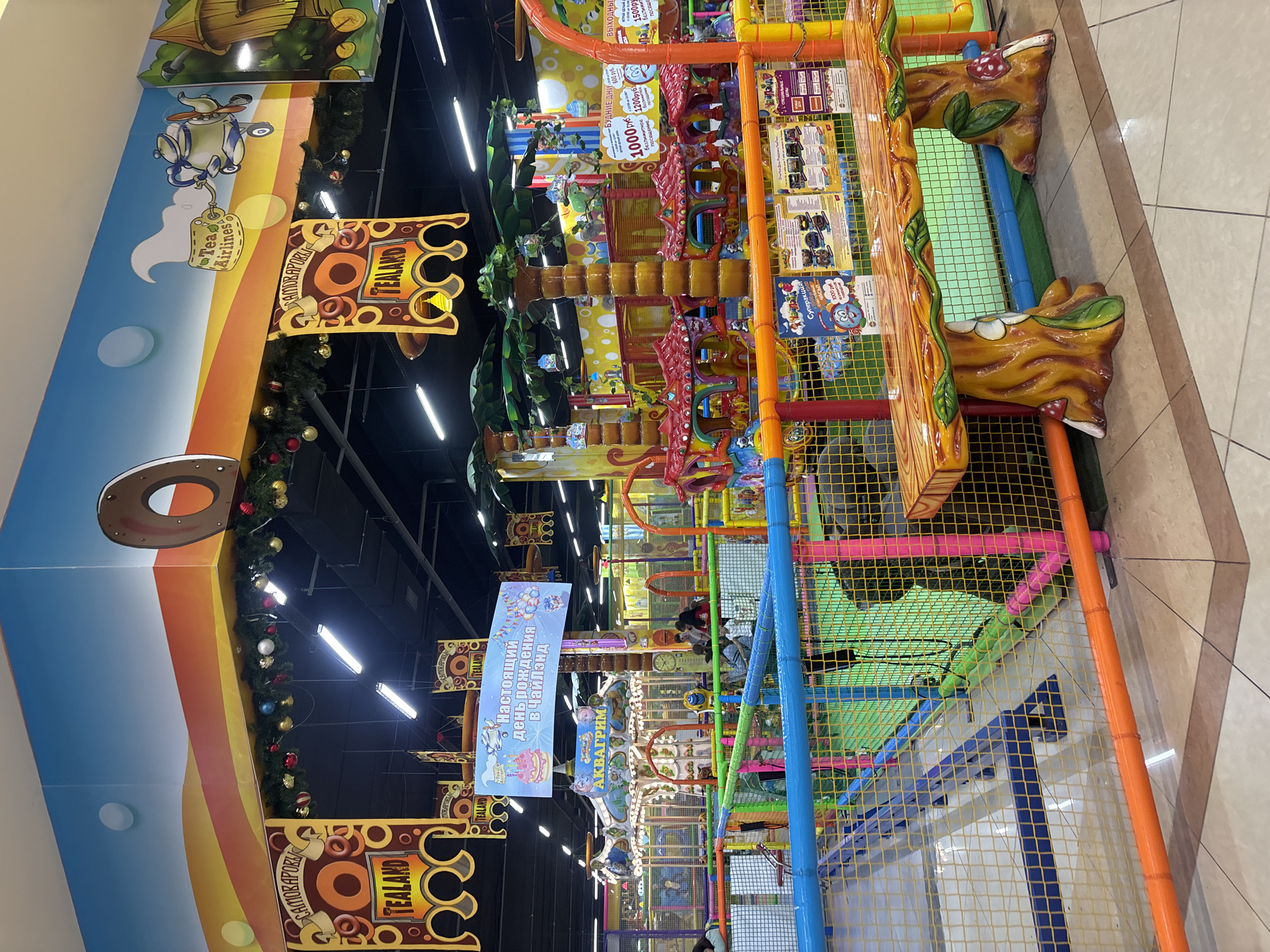 This is a medium shot of a brightly colored indoor children's play area, likely part of a shopping mall or entertainment center in Ivanovo, Russia. The foreground features a large, wooden bench designed to look like a tree trunk with mushrooms and leaves. Behind the bench is a colorful play structure with blue, pink, and orange safety fences. In the middle ground, a carousel with painted horses is visible, along with various climbing structures and slides. The background shows more of the play area, including palm tree-themed decorations and banners. The ceiling is decorated with a sky mural and festive garlands. Several children and adults are scattered throughout the play area, engaged in various activities. The overall atmosphere is lively and recreational.