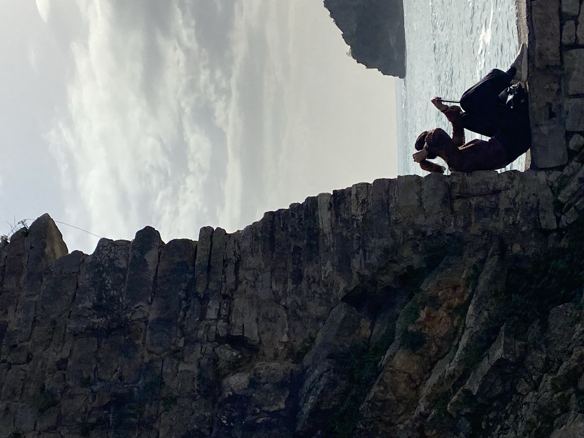 A sideways image captures a person sitting on a stone wall with the sea and sky in the background. The stone wall is made of irregular, dark-colored stones. Green vegetation grows from the cracks and crevices of the wall. A person sits on the edge of the wall, with their legs dangling over the side. They wear dark pants, a tan shirt, and brown shoes. The person is holding up a cell phone, as if taking a picture. Behind the wall is the sea and a rocky islet. The sea appears calm and silvery under a sky with clouds. The image appears to have been taken on a coast near Budva, Montenegro.