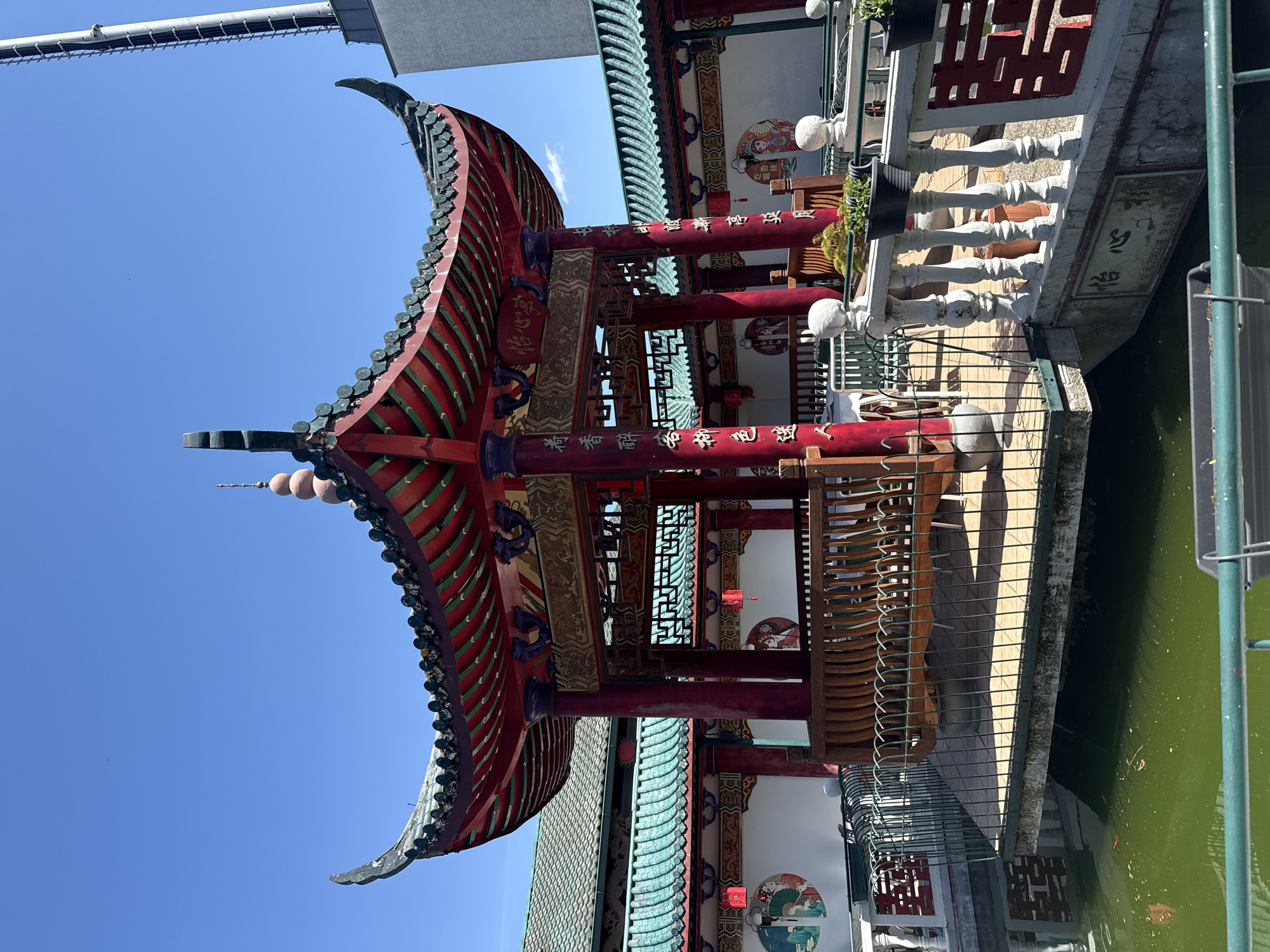 An outdoor, eye-level shot captures a vibrant, traditional East Asian-style pavilion, likely located at the Hainburg an der Donau location provided. The structure features a distinctive curved, multi-tiered roof adorned with red and green/blue tiles, topped with an ornate finial. Below the roof, rich red pillars stand prominently, embellished with intricate golden Chinese characters. Intricate wooden lattice work adds detail to the structure.
The pavilion overlooks a body of water, possibly a pond or canal, which appears green. A white balustrade with ornate spindles separates the pavilion's edge from the water. Potted plants are visible along the balustrade. The background reveals white walls adorned with circular decorative elements featuring traditional Chinese art motifs, such as figures in traditional dress and landscapes. The sky is clear blue, suggesting a sunny day. No people are visible, indicating a serene and tranquil setting.
Visible text on the pillars and a stone plaque includes:
* "怡心亭" (Yí Xīn Tíng), which translates to "Joyful Heart Pavilion."
* "荷香醉客新色迷人," a poetic phrase meaning "Lotus fragrance intoxicates guests, new colors are charming."
* "香" (Xiāng), meaning "Fragrance" or "Incense."
* "吃喝" (Chī Hē), meaning "Eat and Drink."
* "公元2002年9月8日" (September 8, 2002), indicating the date.