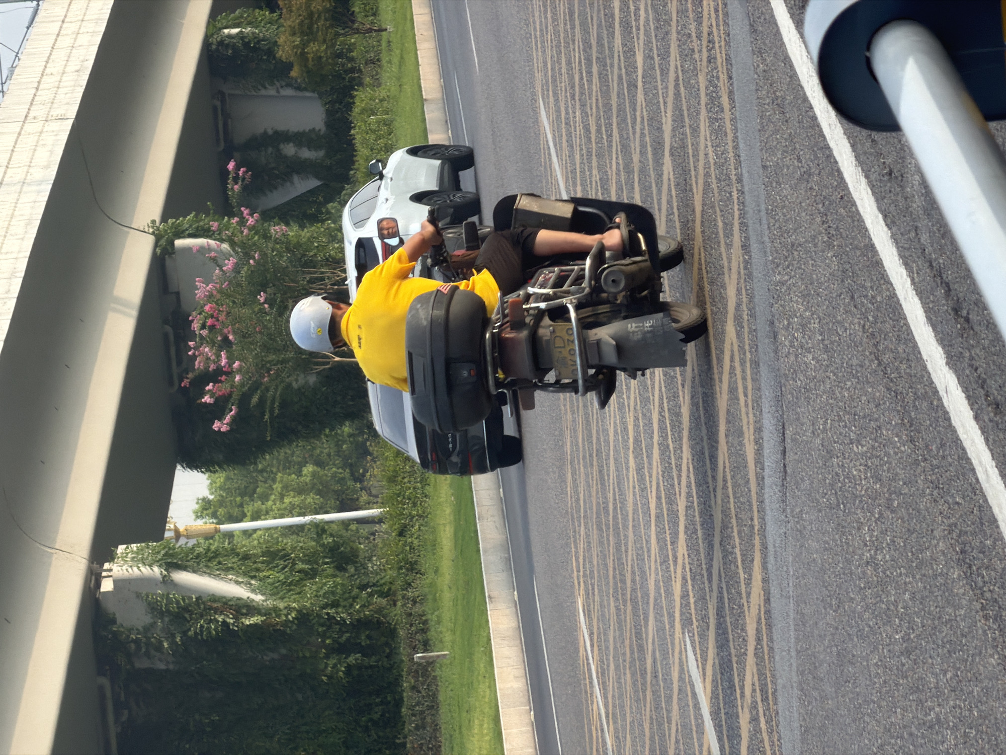 In Jiangjin, China, a person wearing a yellow shirt and a white helmet rides a scooter on a paved road. The scooter has a black storage box attached to the rear. A white SUV is visible to the right of the scooter, and a dark sedan is to the left. The road has yellow and white painted lines. In the background, there is a large concrete overpass covered in green vines and some pink flowering bushes. The sky is overcast.