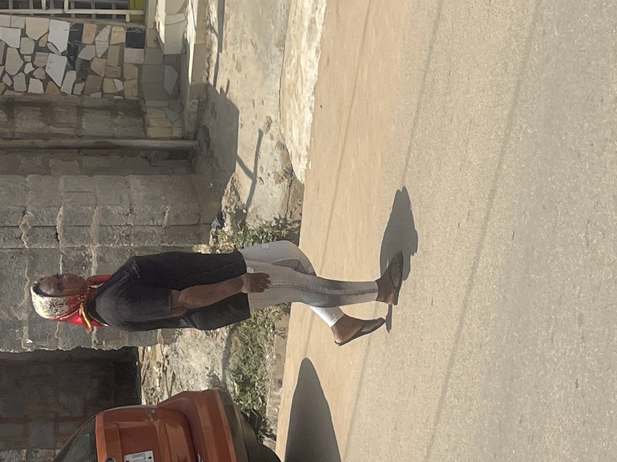 A woman in a black t-shirt, light-colored pants, and a red and white headscarf walks along a paved road, casting a shadow to her left. To her left, an orange vehicle is parked. Behind her, a building is under construction, with walls made of concrete blocks and some sections decorated with irregular stone tiles. Steps lead up to an entrance on the right side of the building. The ground is a mix of concrete and dirt. The scene appears to be in Abuja, Nigeria.