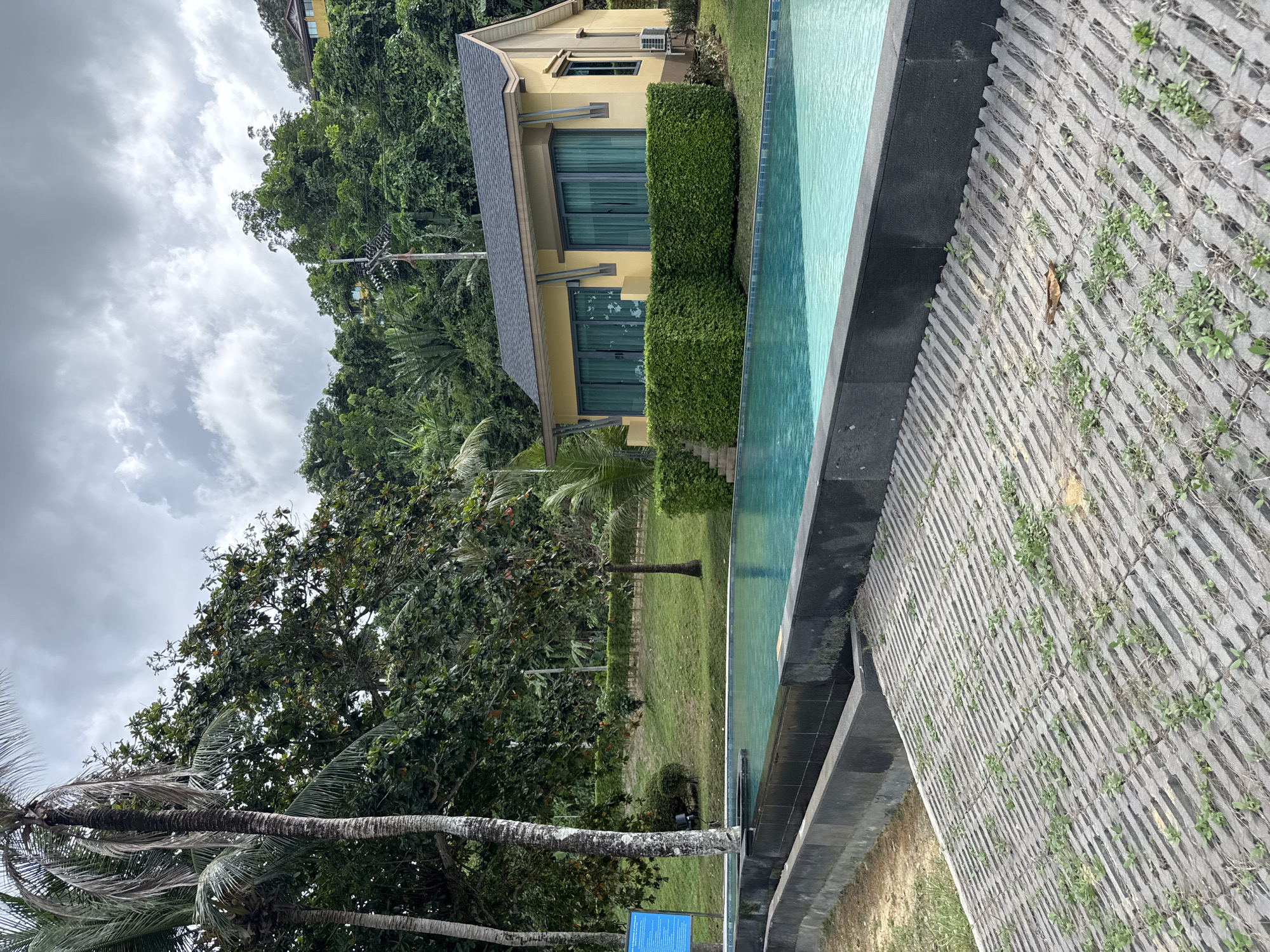 This is a horizontal, eye-level, medium shot of an infinity pool in front of a yellow villa. The pool is filled with clear, turquoise water and is bordered by dark gray stone. Beyond the pool, to the left, is a large palm tree with fronds that obscure the cloudy, gray sky. A blue sign is attached to a post to the left of the palm tree. To the right of the pool is a sloped, gray concrete area with a ribbed texture, and patches of green weeds are growing between the ribs. In the background, a small, yellow villa with a dark gray roof is nestled into a lush, green hillside. The villa has large windows with blue curtains and an outdoor air conditioning unit attached to the right side. The overall scene suggests a resort or private residence in a tropical location. The location is Pa Khlok, Thailand.