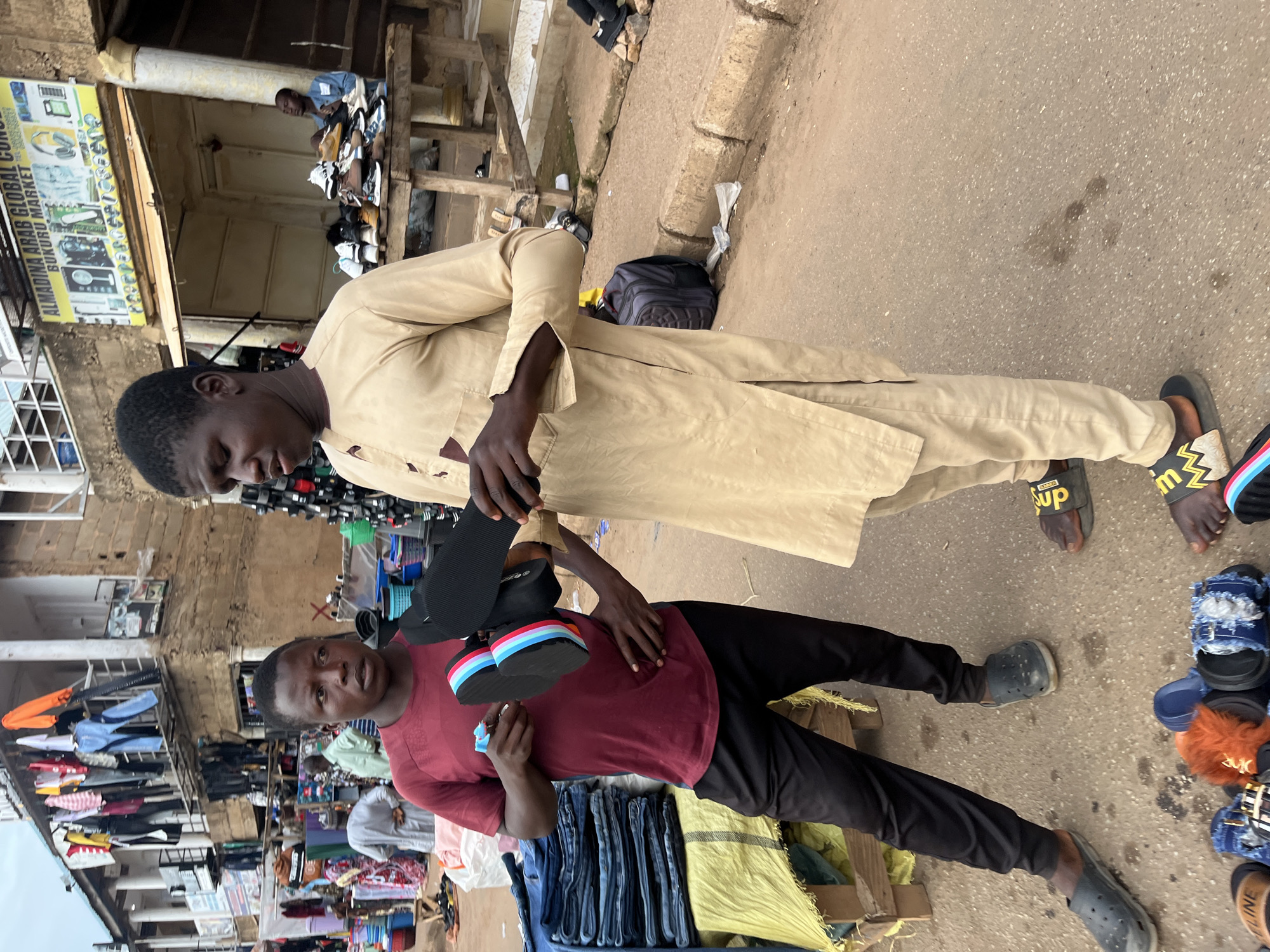 The image shows two young men in an outdoor market setting in Bukuru, Nigeria. One man, wearing a red t-shirt and black pants, holds a pair of colorful flip-flops in his left hand and appears to be presenting them or showing them to the other man. The second man, dressed in a tan traditional garment, is holding a black flip-flop and looking at it. Several stacks of blue jeans are visible on a wooden stand to the left of the first man. In the background, there are market stalls with clothing and merchandise displayed, as well as a sign for "ALMADINA ARAB GLOBAL CONCERN BUKURU MARKET." Various footwear, including sandals and shoes, are displayed on tables and on the ground in front of the men. The ground is paved with asphalt.