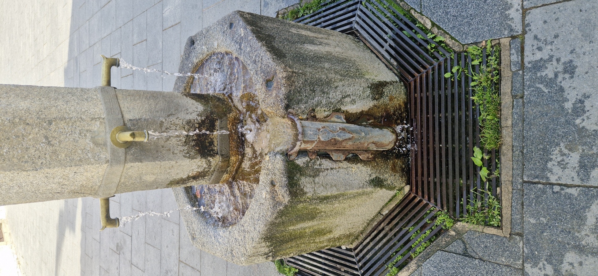 This is a high-angle, close-up, full-shot photograph of an old, octagonal stone fountain with two spigots made of brass. Water is flowing from both spigots, with one stream landing in the basin of the fountain and the other flowing directly into a metal grate below. The fountain has a weathered appearance, with green moss and dark stains on the stone. The basin is filled with water, and a metal spout extends from the front of the fountain, directing overflow into the grate. The grate is part of a drainage system surrounding the fountain and is partially covered by grass and weeds. The fountain is situated on a paved area, possibly a public square or walkway, with cobblestone-like tiles visible. The lighting suggests it is daytime, with shadows cast on the ground.