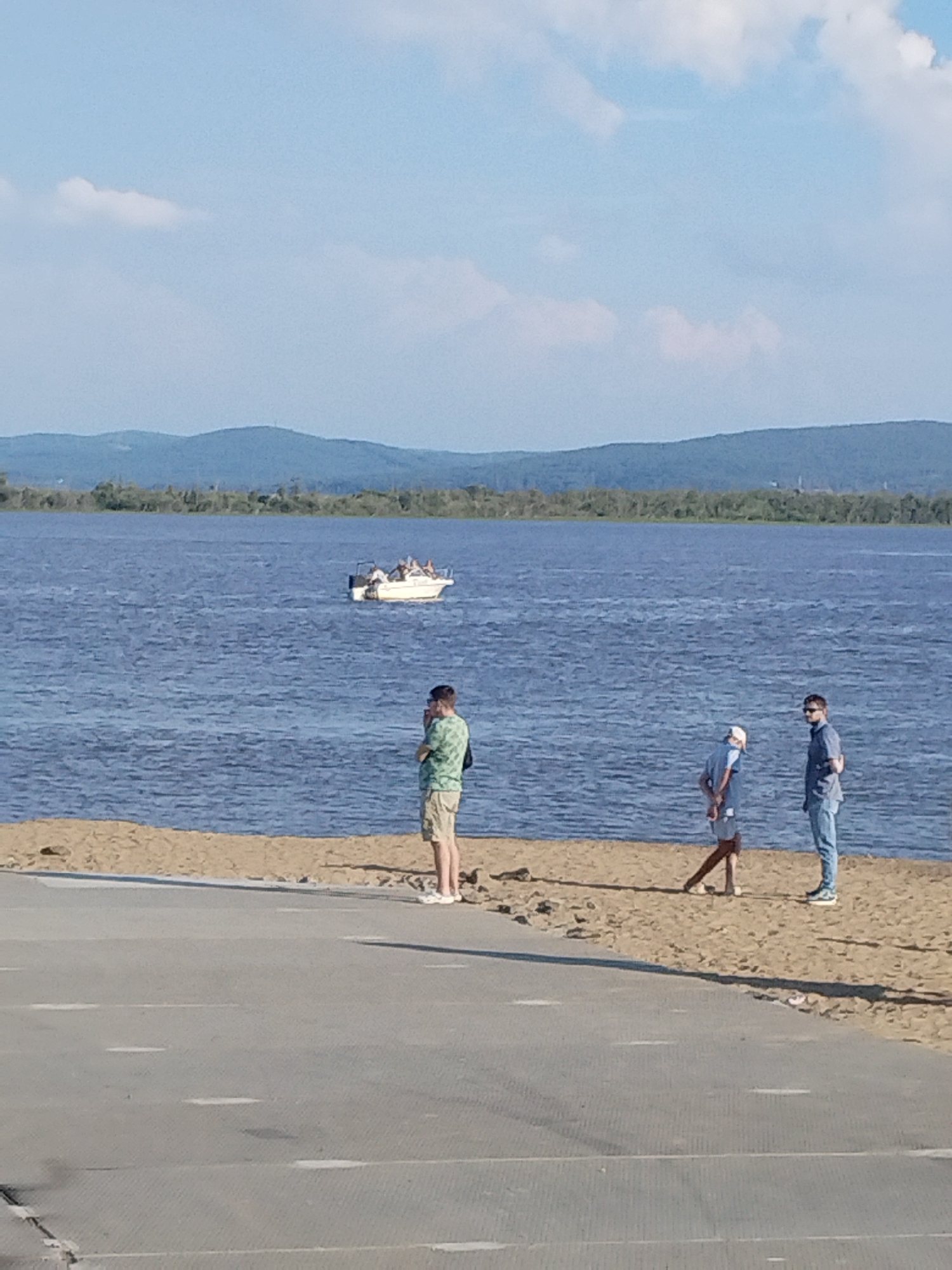 The image depicts a wide, calm body of water, likely a river, under a bright, partly cloudy sky. In the distance, rolling, tree-covered hills form the horizon. On the water, a small white boat with several passengers is visible. The foreground features a sandy beach, where three individuals are standing. To the left, a person in a green patterned shirt and khaki shorts stands facing away from the camera. To the right, two individuals are standing closer to the water. One, wearing a light blue shirt and white cap, faces left. The other, in a dark blue shirt and jeans, faces right. A paved surface, possibly a walkway or road, occupies the very bottom of the frame. The scene is consistent with a recreational area along the Amur River within the city of Komsomolsk-on-Amur, Russia, likely during daylight hours in warmer weather.