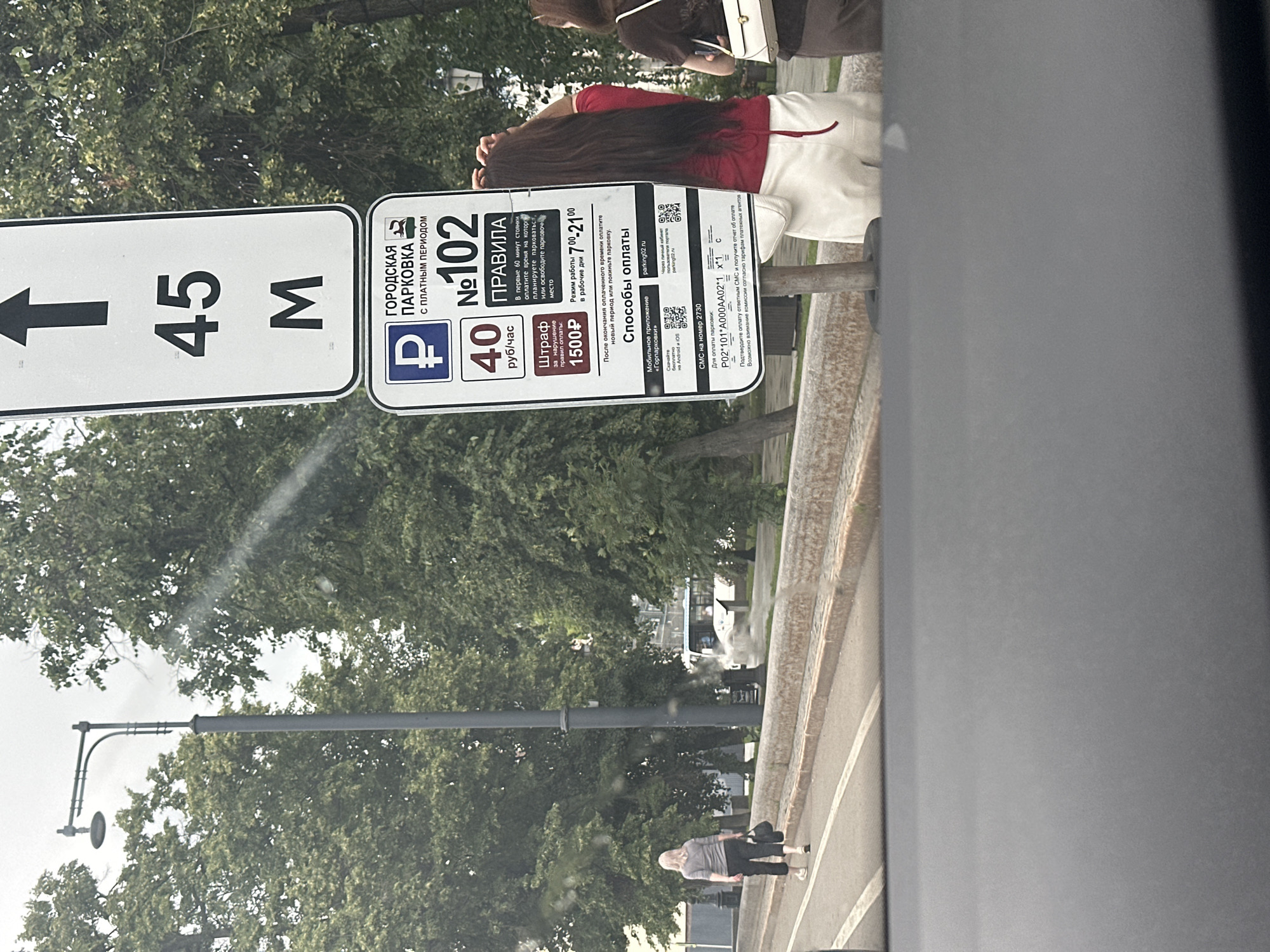 In Ufa, Russia, a parking sign is prominently displayed. The sign indicates a parking zone with a paid period and a numbering system, with "№102" clearly visible. It also specifies a fine of "1500₽" for rule violations. The sign provides information on payment methods, including a mobile application and SMS services, and lists operating hours from "7⁰⁰-21⁰⁰". An arrow pointing upwards with "45 M" indicates a 45-meter distance, likely related to parking regulations. A woman with long dark hair, wearing a red top and white pants, stands near the sign, adjusting her hair. Another woman in the background is walking away, carrying a bag. The scene is set outdoors, with trees and a street visible. The overall environment suggests a public urban space, possibly a street or park with designated parking.