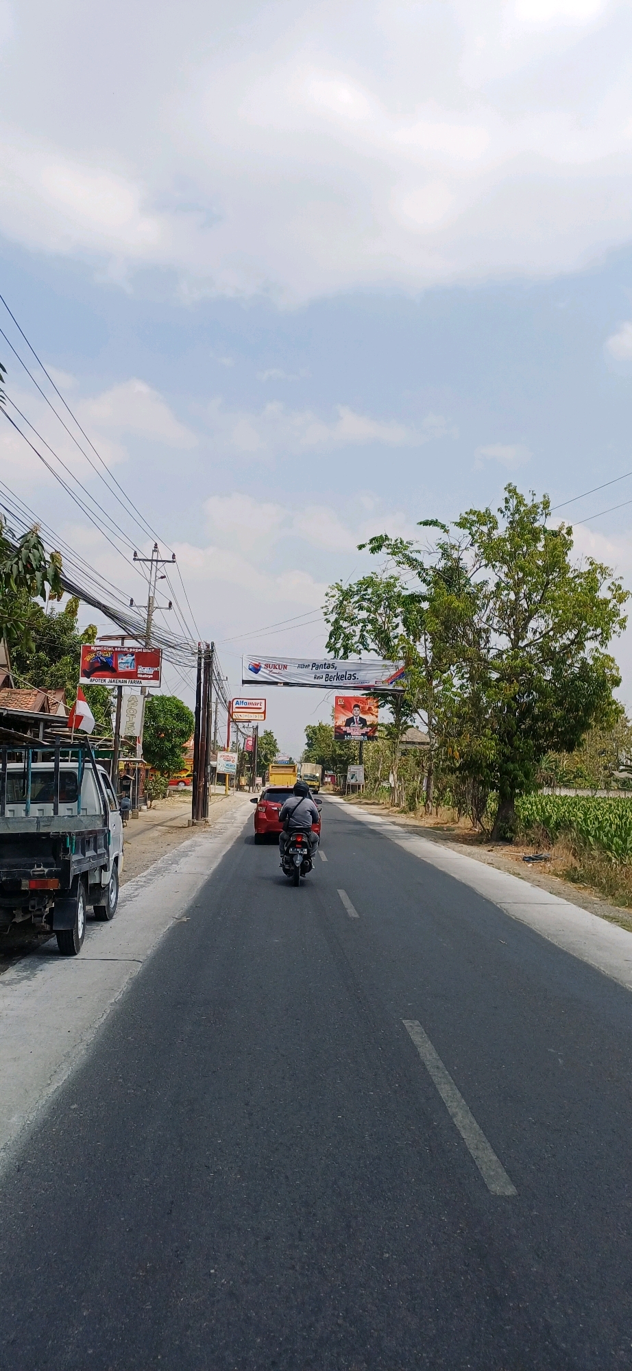 This is a vertical, eye-level, daytime, outdoor shot captured on a road in Jakenan, Indonesia. The perspective is from behind a motorcycle rider. The road is asphalt, with a white dashed line marking the center. On the left side of the road, there's a gray flatbed truck. Further down the road on the left, there are buildings, utility poles with numerous wires, and trees. A billboard for "APOTEK JAKENAN FARMA" is visible. On the right side of the road, there's a large tree and a field of crops. Overhead, a banner reads "SUKUN Pantas, Rasa Berkelas." Below this banner, there's a red billboard featuring a man in a suit, likely a political advertisement, and an Alfamart sign. A red car is driving ahead of the motorcycle. The sky is partly cloudy and light blue.