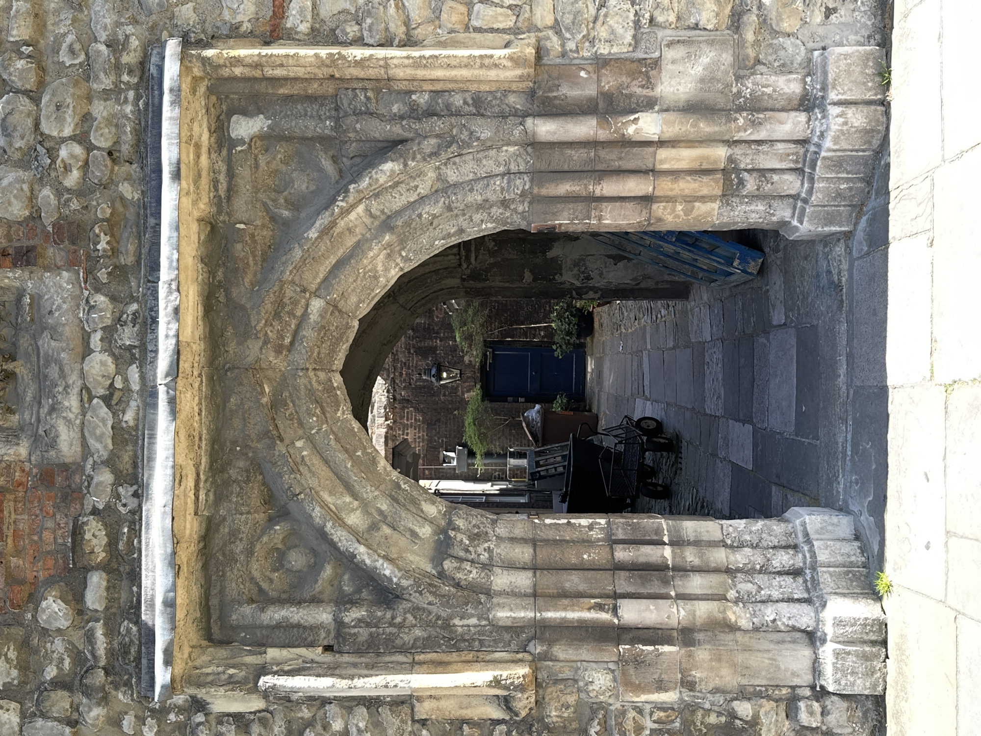 The image presents a stone archway, possibly the remains of a historic structure, constructed with aged stone blocks. The archway leads into a narrow passageway or courtyard paved with stone. Within the courtyard, a black trolley and other objects are visible. A blue door is at the end of the passage. Vegetation is growing near the entrance. The arch is a part of the Tower of London in the United Kingdom. The image is taken at eye-level.