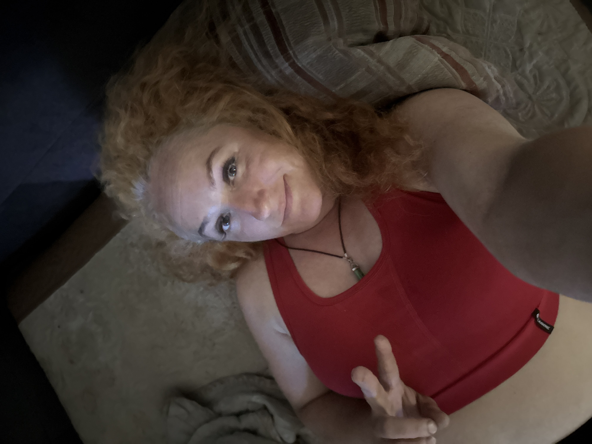 A woman with curly red hair and light skin lies on her back, looking up at the camera and making a peace sign with her left hand. She is wearing a red tank top and a necklace with a green pendant. The background appears to be a bed with striped pillows. There is no discernible location in the city of Chaikovsky, Russia.
