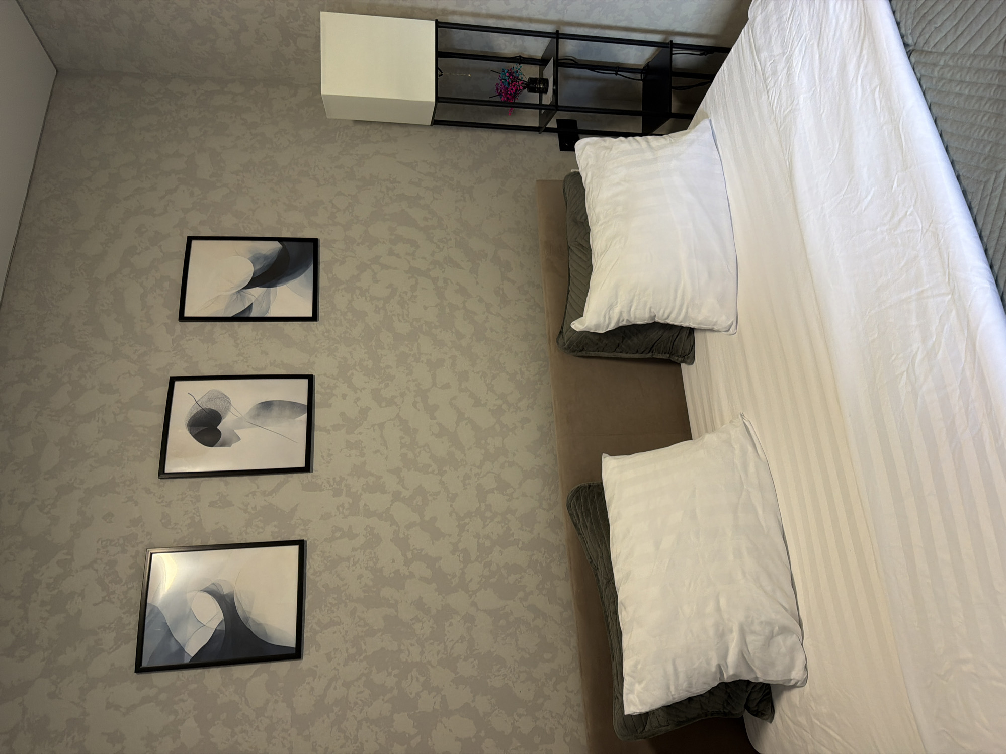 The image shows a bedroom with a bed, a floor lamp, and three framed artworks on the wall. The bed is made with white striped sheets and two pillows, one dark green and one white. The floor lamp is tall and has a white square shade. The artworks on the wall are abstract and feature shades of blue and gray. The wall has a textured gray wallpaper. The room appears to be clean and well-lit, with a neutral color palette.