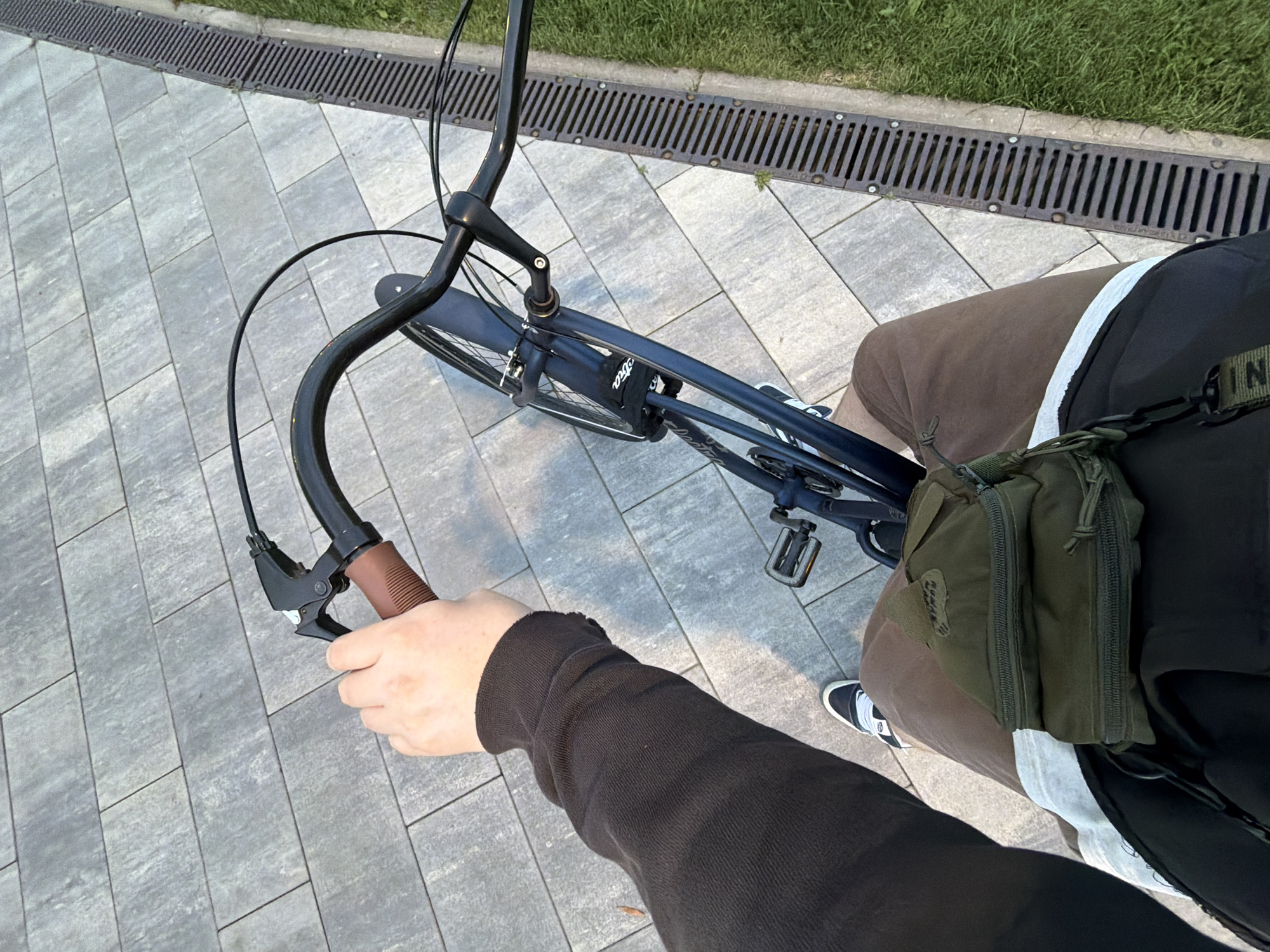 A first-person perspective shot shows a person riding a bicycle on a paved path in Khimki, Russia. The cyclist is wearing a brown long-sleeved shirt, brown shorts, and white sneakers with black accents. A dark green utility pouch is slung across their waist. The bicycle is dark blue with black handlebars and pedals. The path is made of rectangular grey paving stones, and to the right, a metal grate runs alongside a patch of green grass. The lighting suggests it is daytime, possibly late afternoon or early evening.