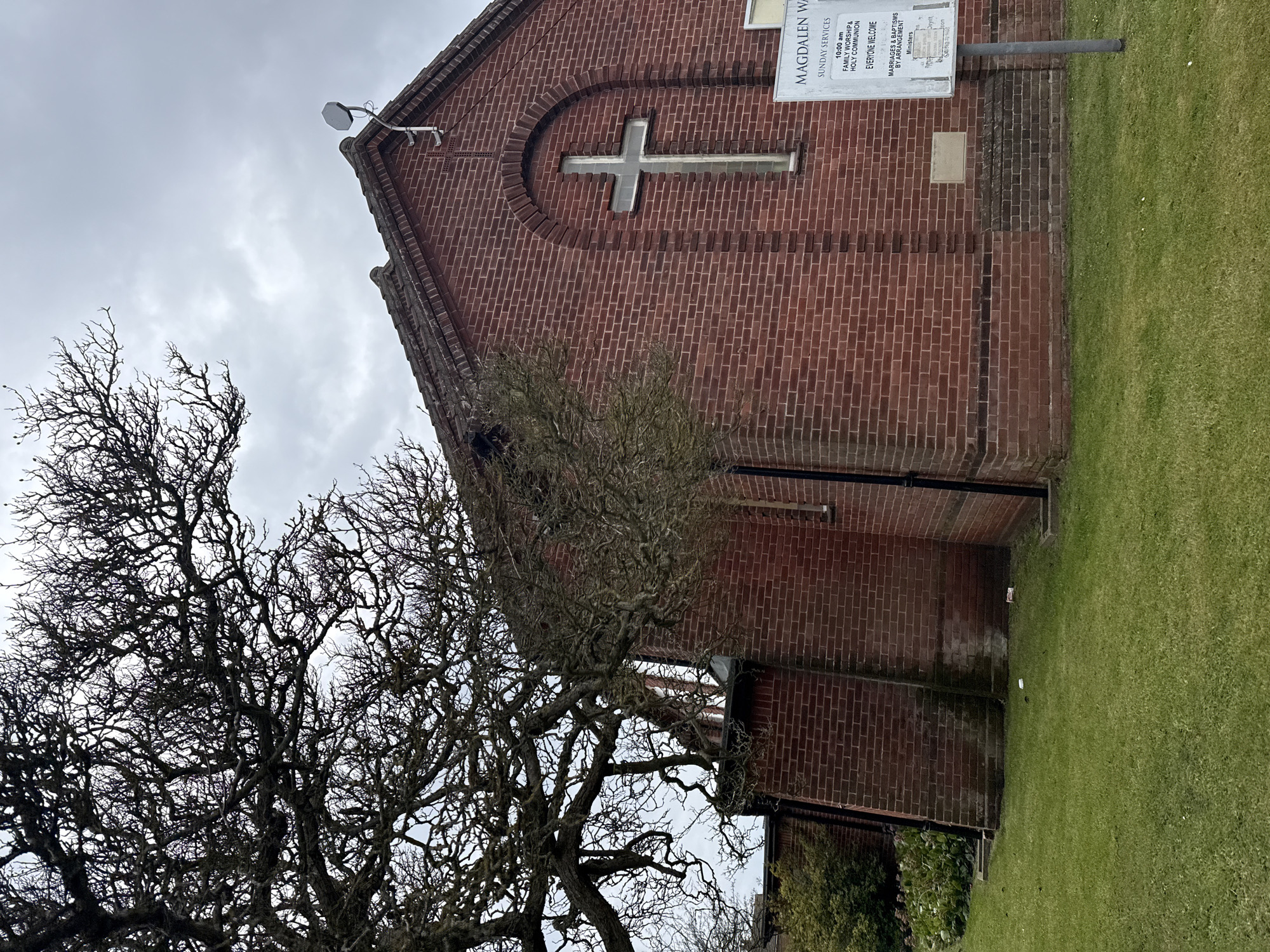The image captures the exterior of Magdalen Way Methodist Church in Gorleston-on-Sea, United Kingdom. The building is constructed of red brick and features a large brick-framed cross above a window. A sign on the side provides details of Sunday services and contact information. A large, bare tree dominates the left side of the frame, contrasting with the green lawn adjacent to the church. The sky is overcast, suggesting a cloudy day. A satellite dish is mounted at the top of the roof.