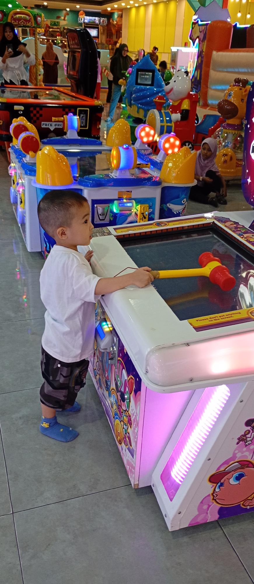 A young boy, wearing a white t-shirt, camouflage shorts, and blue socks with yellow cartoon characters, stands at a colorful arcade game. He holds a yellow and red toy hammer, poised to strike the game's surface. The arcade machine is adorned with vibrant cartoon graphics and lit by pink and blue lights. In the background, other arcade games and people are visible, including women in headscarves. The scene takes place indoors on a tiled floor, suggesting an amusement center or entertainment venue in the city of Probolinggo, Indonesia.