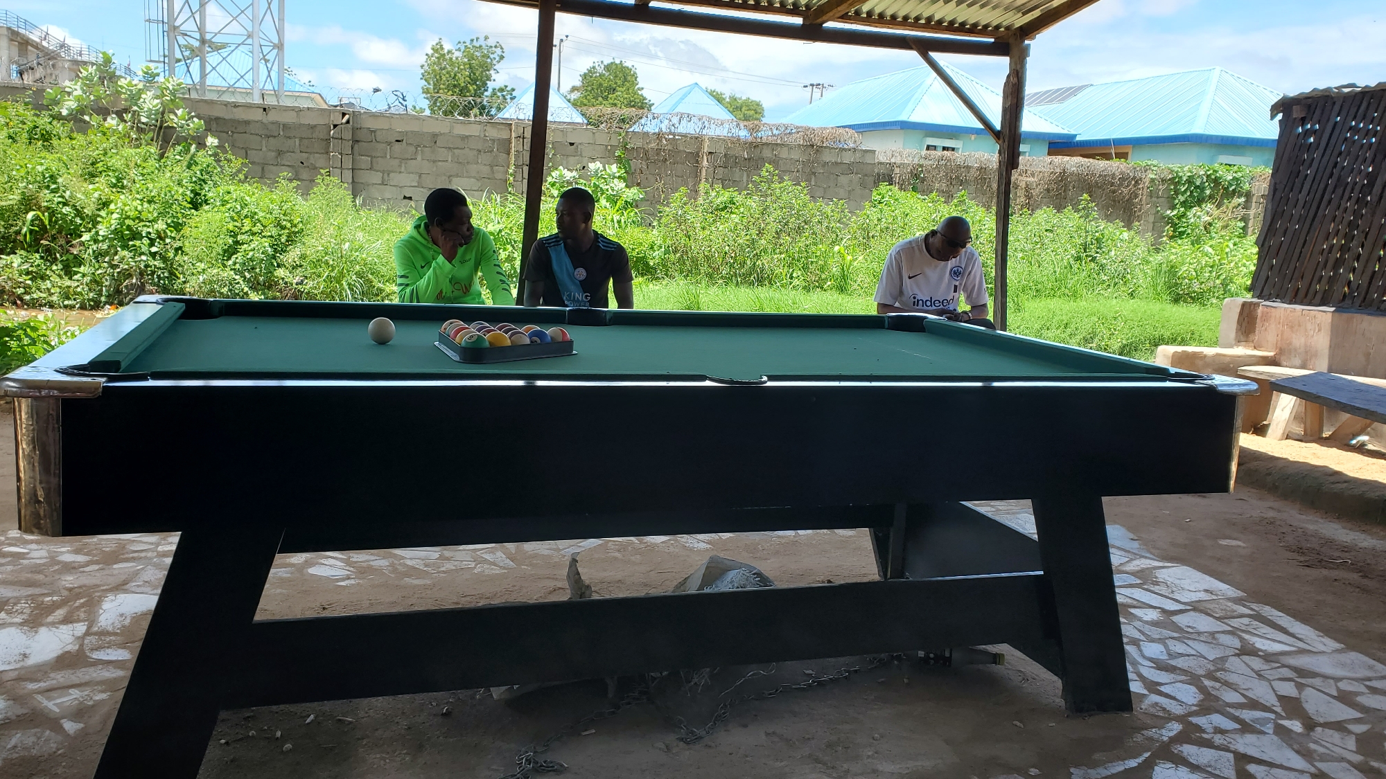 In Jalingo, Nigeria, under a corrugated metal roof supported by wooden posts, three men stand around a dark green pool table. The table is set with a full rack of billiard balls and a single white cue ball positioned near the center. The man on the left wears a bright green long-sleeved shirt and leans forward, his hand near his face. The man in the middle wears a dark t-shirt with a graphic print. The man on the right wears a white t-shirt with a logo that reads "indeed." Behind the pool table, a low concrete wall topped with barbed wire separates the area from a dense growth of green vegetation. In the background, several buildings with blue, corrugated metal roofs are visible under a bright, partly cloudy sky. The ground in the foreground is a mixture of concrete tiles and dirt.