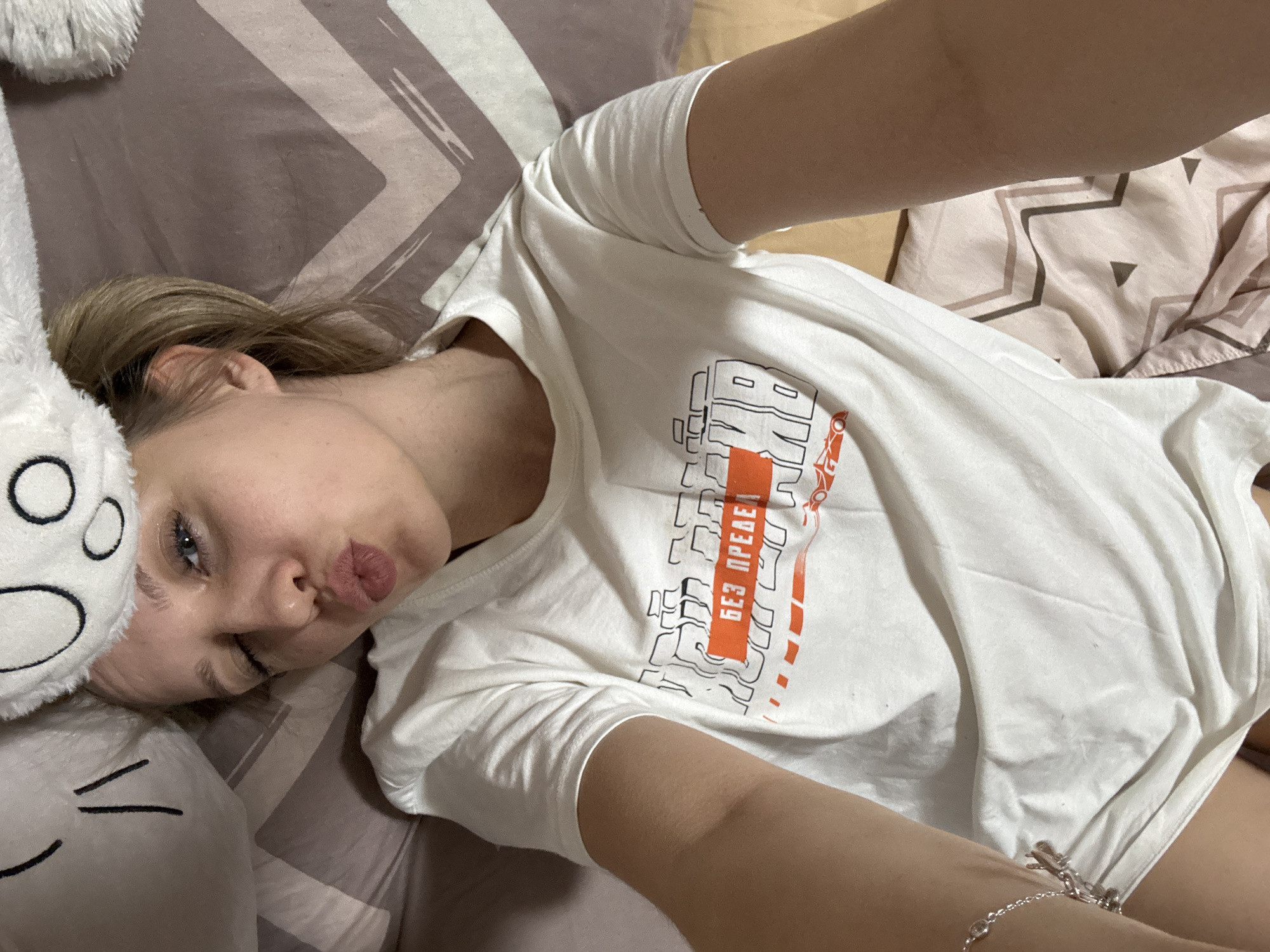 A woman is lying on a bed. She is wearing a white t-shirt with an orange stripe and Cyrillic text. The woman has fair skin, blue eyes, and light brown hair pulled back. She is making a kissing face. A white plush toy is next to her head. The bedding has a brown and white geometric pattern. The location is Unknown, Unknown.