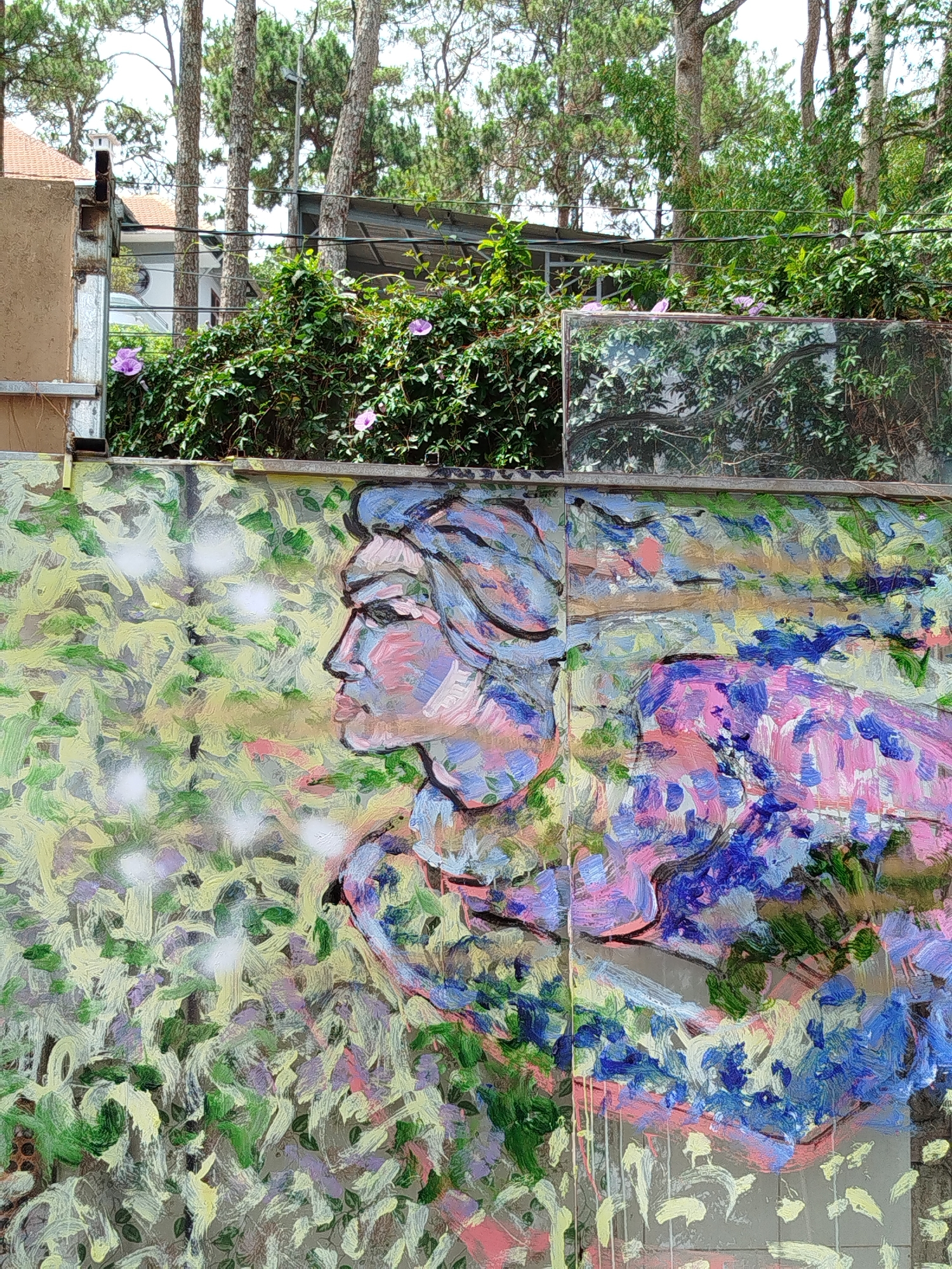 A mural in Da Lat, Vietnam, depicts a stylized profile of a person against a backdrop of green foliage and dappled light. The mural uses visible brushstrokes and a palette of blues, pinks, greens, and yellows. The subject's face is turned to the left. Above the mural, vegetation with purple flowers grows along a wall. A glass pane is visible, reflecting trees and sky. Beyond the wall, a building with a metal roof is partially visible among tall trees.