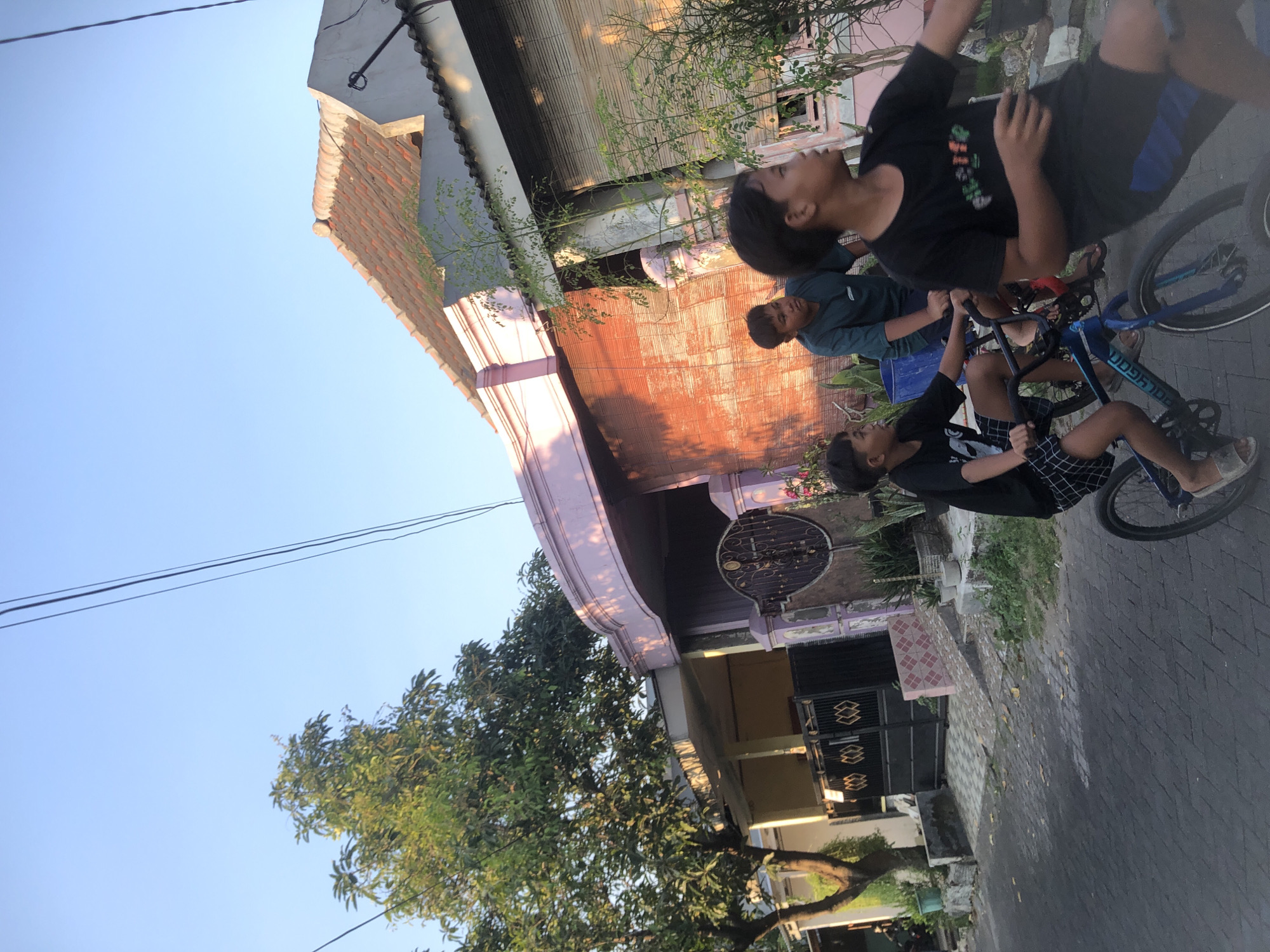 The image depicts a street scene in Kalisampurno, Indonesia, bathed in the warm light of the late afternoon sun. Three young males are actively engaged in riding bicycles on a paved road. The foreground is dominated by a boy in a black t-shirt and checkered shorts, positioned on a blue bicycle, with his right leg extended as if pedaling. To his left, another boy in a black t-shirt and plaid shorts is also on a bicycle, his gaze directed forward. Behind them, a third boy, wearing a teal shirt, is seated on a bicycle, looking towards the left. The environment includes residential buildings with tiled roofs and walls adorned with decorative elements. A large tree with green foliage stands to the left of the houses, casting shadows on the scene. Power lines are visible crisscrossing the clear blue sky. The overall atmosphere suggests a casual, everyday activity within a residential neighborhood.