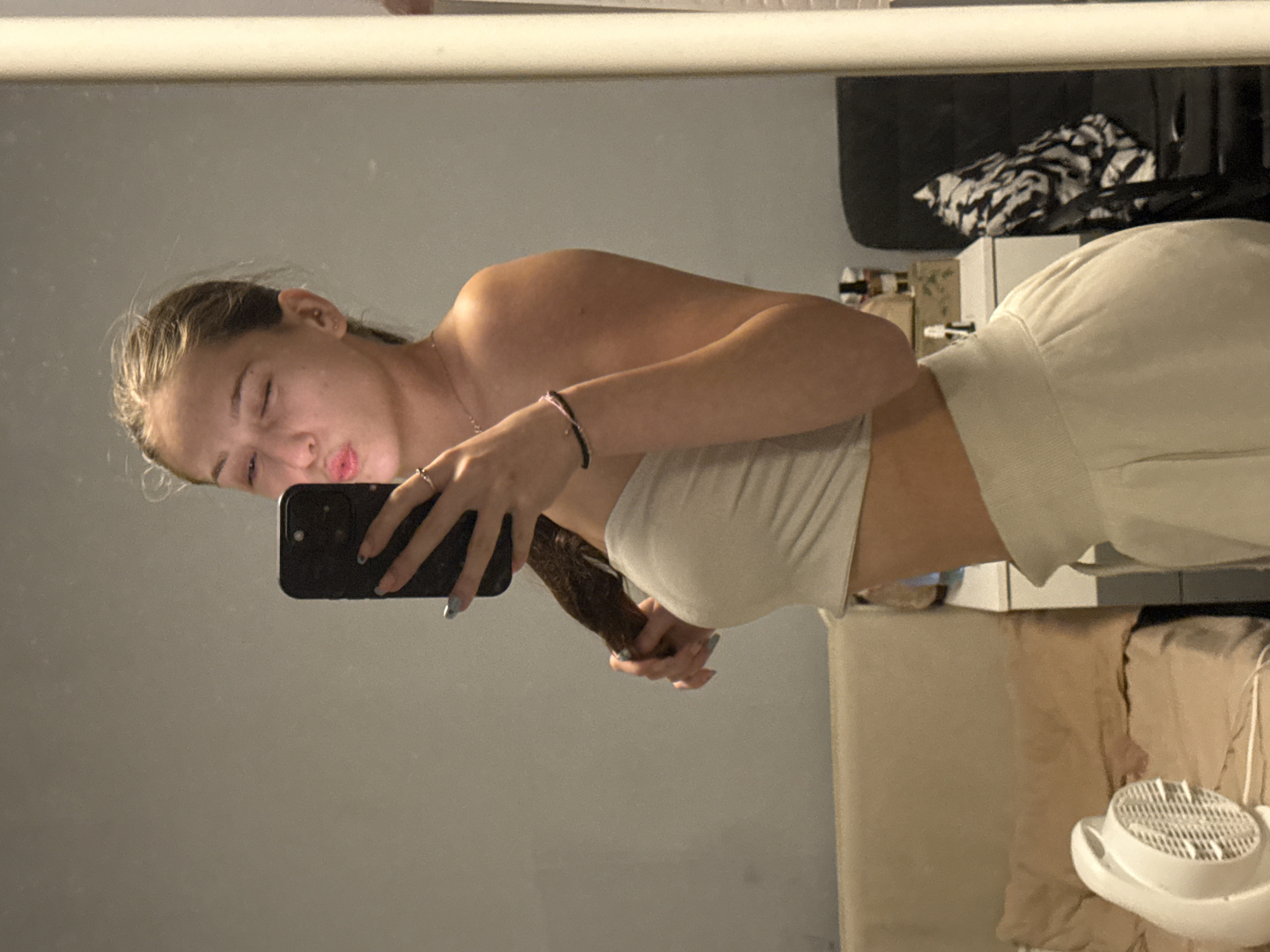 A young woman with blonde hair tied back is taking a mirror selfie in what appears to be a bedroom. She is wearing a beige tube top and matching sweatpants. Her right arm is holding a black smartphone, and her left hand is holding a section of her hair. She is winking and puckering her lips. In the background, a bed with beige bedding and a pillow is visible, as well as a white fan on the left. A bedside table with some items on top is on the right, and a black chair with a black and white patterned item draped over it is reflected in the mirror. The lighting is dim, suggesting it is nighttime or the room is dimly lit. There are no identifiable landmarks or indications to suggest the image was taken in Sofia, Bulgaria.