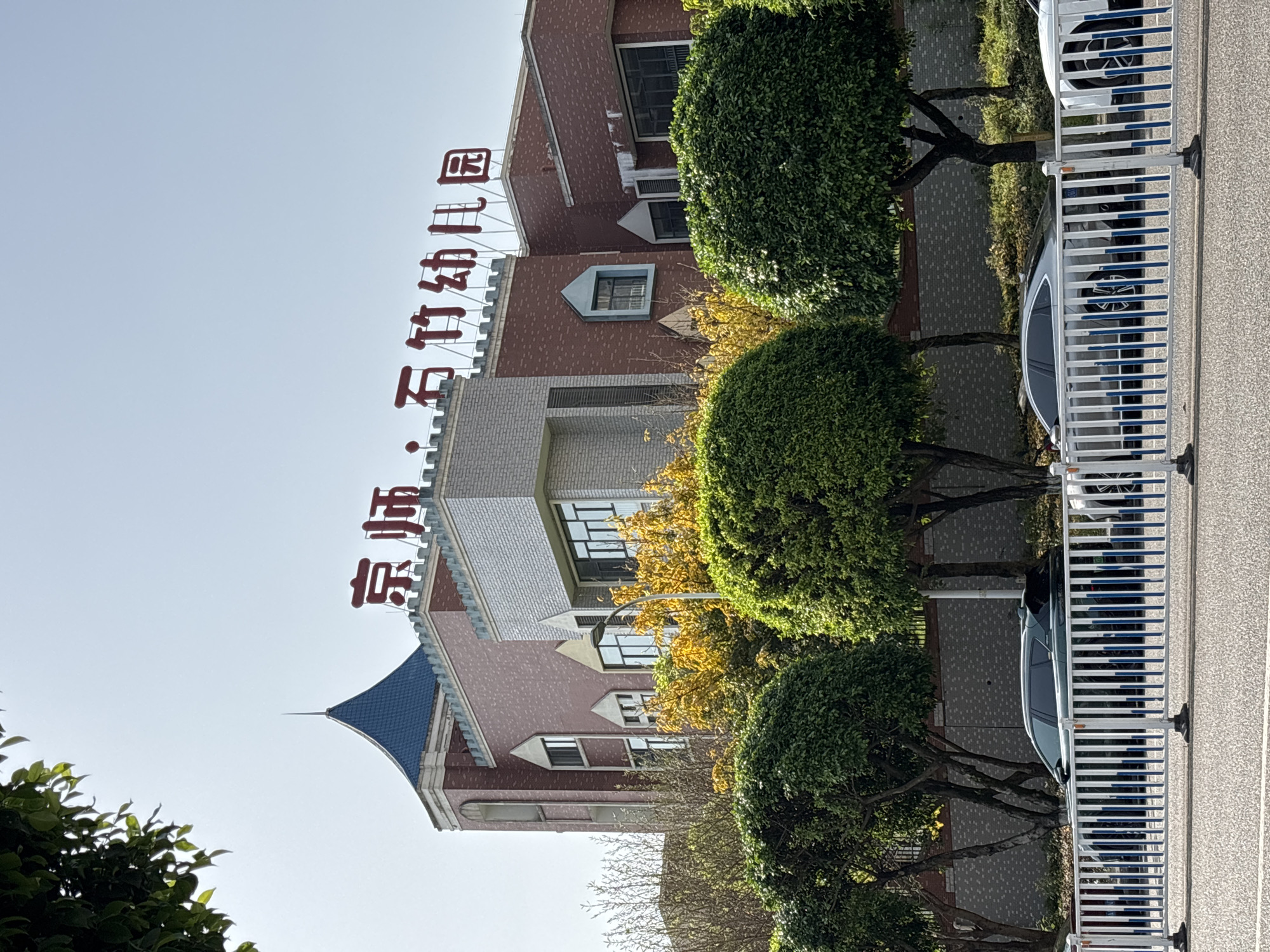 This media file captures a bright, sunny daytime scene of a building, likely a kindergarten, in Jiangjin, China. The image is taken from a street-level perspective, looking towards the building.
The main subject is a multi-story building with a distinctive architectural style. Its facade is primarily composed of red brick-like material on the upper sections, contrasting with lighter-colored brick or tile on the lower parts. The roofline features small, castle-like crenellations. A prominent blue, conical-shaped roof section with a spire is visible on the left side of the building. Several windows with dark frames are scattered across its face.
Above the building, large red Chinese characters are mounted, spelling out "京师石竹幼儿园" (Jīngshī Shízhú Yòuéryuán), which translates to "Jingshi Shizhu Kindergarten."
In front of the building, a row of well-trimmed, spherical green trees lines what appears to be a sidewalk or median. Some trees exhibit yellowing leaves, suggesting autumn. To the right, a white fence with vertical blue stripes separates the pavement from the street. Several cars, predominantly white and dark grey, are parked on the street beyond the fence.
The sky is clear and a light blue, indicating fair weather. The lighting is bright and direct, typical of daylight hours. The scene appears static with no discernible activity or people present.