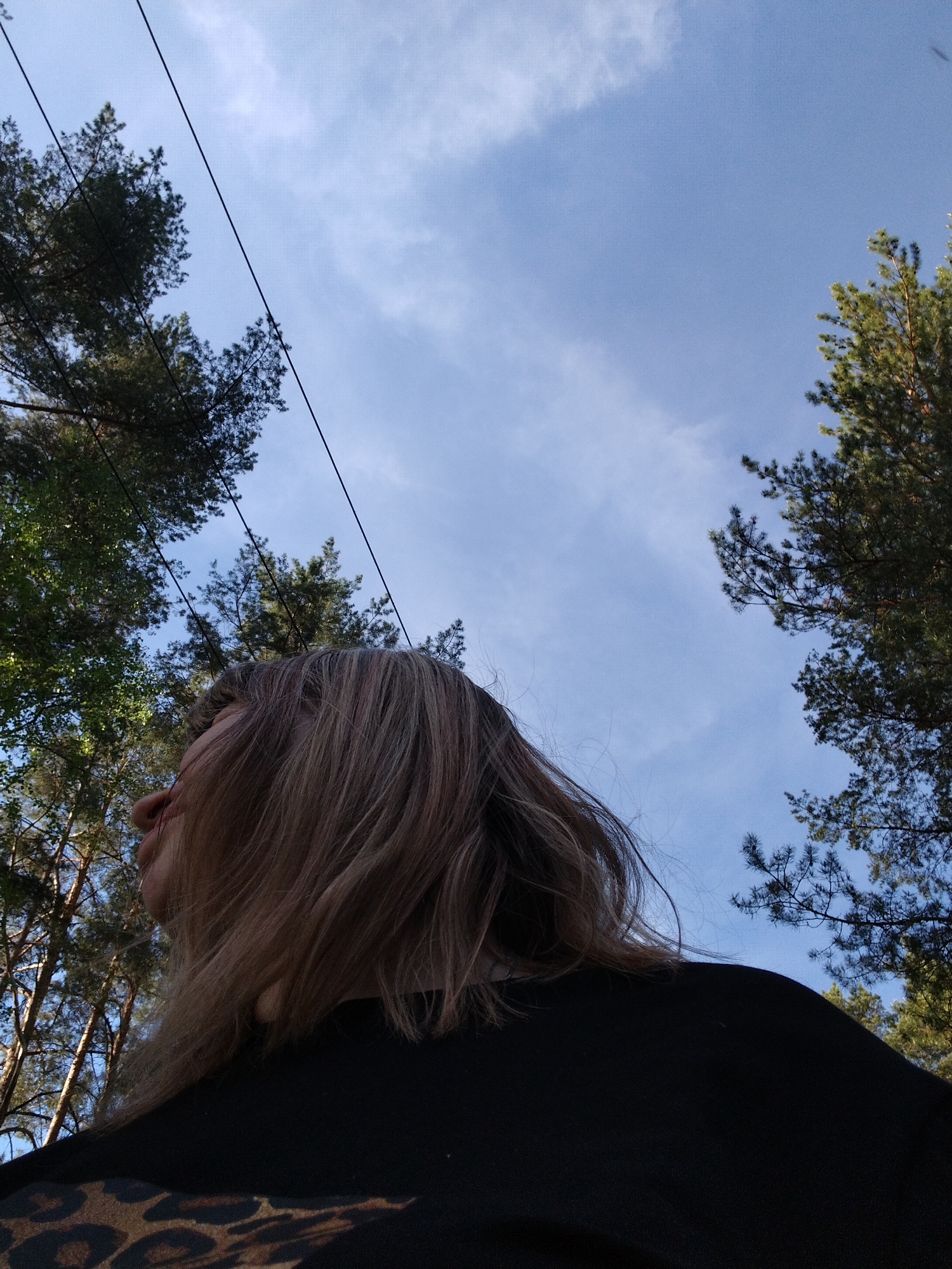 The photograph shows a woman looking upwards in Stroitel', Russia. She has shoulder-length, light brown hair, and is wearing a black shirt with a leopard print pattern. The view is angled upwards, revealing a light blue sky with wispy white clouds. Tall trees frame the sky on both sides, with power lines visible against the sky in the upper left corner of the frame. The light suggests it's daytime.