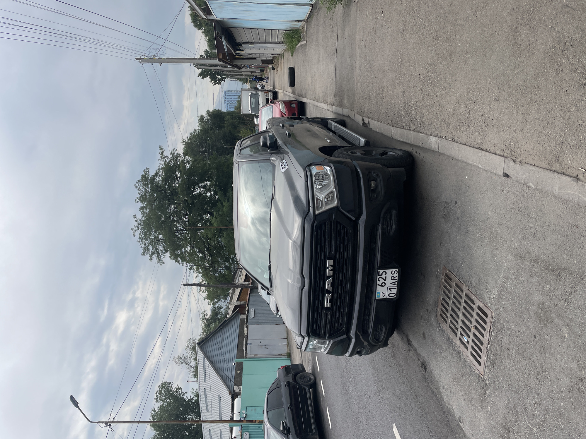 A black Ram pickup truck is parked on the side of a paved road in Almaty, Kazakhstan. The truck is facing forward, slightly angled towards the right side of the frame. A license plate with "KZ 625 01ARS" is visible on the front bumper. To the left of the truck, a black sedan is also parked on the road. The road has a white line marking the edge of the lane. In the foreground, a metal grate for a storm drain is set into the asphalt. To the right of the truck, a sidewalk runs alongside the road, with several parked cars and a white truck visible further down. The sidewalk is bordered by a fence and buildings. The sky is overcast with scattered clouds. Trees are visible behind the buildings. The scene appears to be in a residential or commercial area.