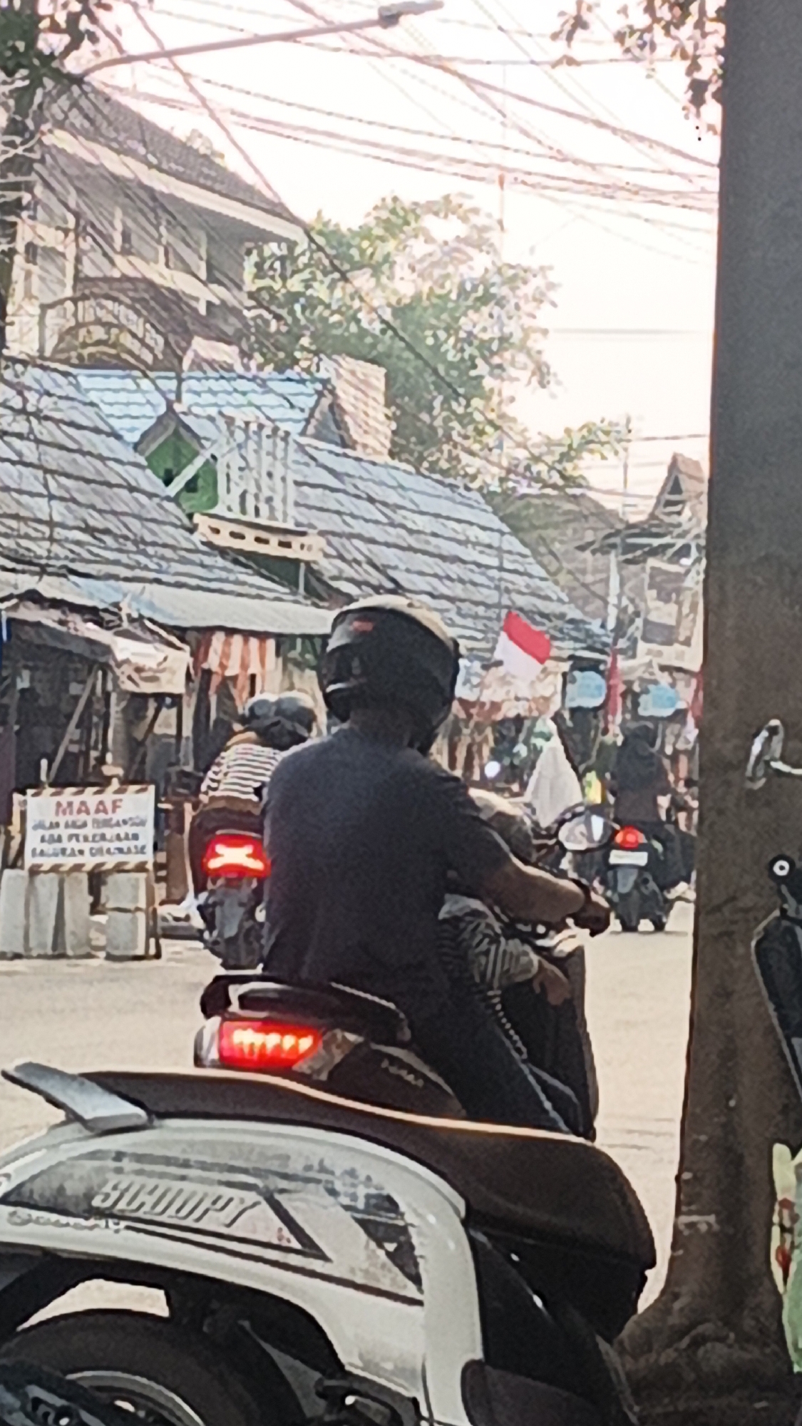 A medium shot captures a street scene in Banjarmasin, Indonesia, with a focus on a person riding a motorcycle. The person, wearing a dark shirt and a black helmet, is positioned in the foreground, obscuring their face. They are on a white and silver scooter, with the model name "SCOOPY" visible on the side. The motorcycle's brake light is illuminated red. Behind the rider, another motorcycle is partially visible, also with a rider wearing a helmet and a striped shirt.
The background reveals a bustling street with several buildings, some with corrugated metal roofs and others with more traditional architecture. A prominent sign in the mid-ground reads "MAAF" (meaning "Sorry" in Indonesian) followed by text indicating road work or closure due to "PEKERJAAN" (work). A small Indonesian flag is visible flying from a pole further down the street. The environment appears somewhat cluttered with various structures and overhead electrical wires, suggesting a densely populated urban area. The lighting indicates it is daytime, possibly late afternoon, with a soft, diffused light.