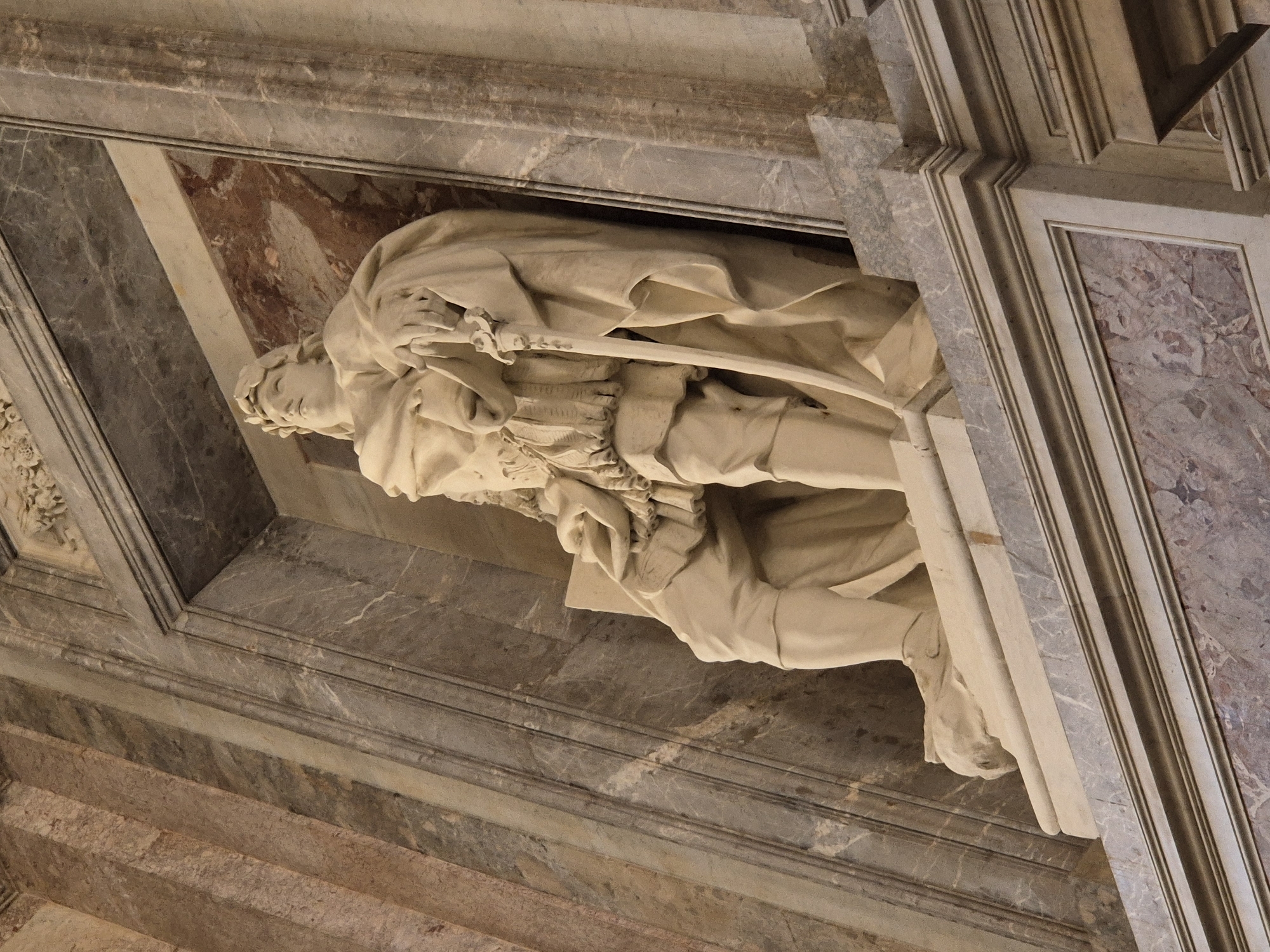 This media file showcases a detail of a sculpture of a person, possibly a man in historical attire, lying down or in a recumbent pose. The sculpture appears to be made of light-colored stone or marble. The figure is draped in flowing robes, with a sword held across its body. The head of the sculpture is visible, tilted downwards with closed eyes, suggesting a state of rest, sleep, or perhaps death. The detailed rendering of the fabric folds and the facial features are notable.
The sculpture is set within an architectural context, framed by marble elements. The surrounding stonework features intricate detailing and veined marble in shades of grey, white, and pinkish-brown. The texture of the marble suggests a grand, possibly historical, indoor setting, consistent with a tomb, monument, or a richly decorated architectural space. The lighting in the scene is subdued, casting soft shadows that accentuate the sculpture's form and the surrounding architectural details. There is no visible text in the image. The orientation of the image is landscape and the photo appears to be taken from a low angle looking upwards. Given the location context of Caserta, Italy, this sculpture could be part of the Royal Palace of Caserta or a related historical site, a location known for its opulent architecture and art.
