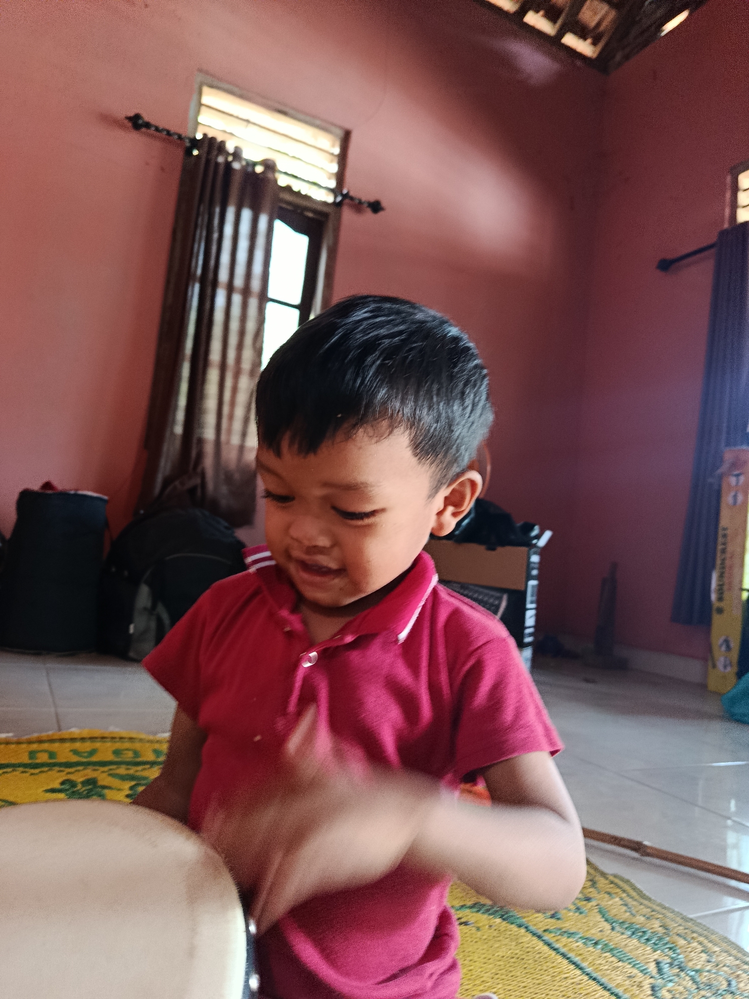 A young boy with dark hair and wearing a red polo shirt is sitting down and playing a drum. He is looking down at the drum with a smile. He is sitting on a yellow mat with a green pattern. The room has pink walls and a wooden ceiling. There are two windows with dark curtains. There are also two black bags in the background. The image appears to be taken indoors.